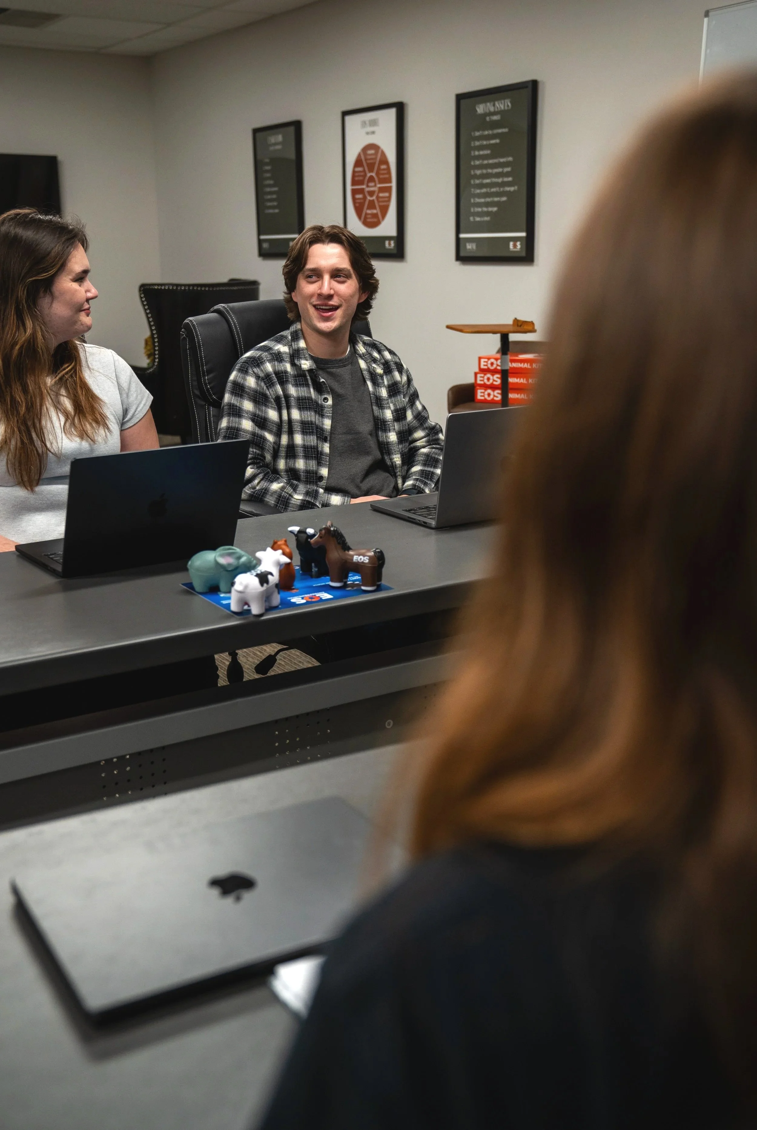 People in a meeting room engaged in conversation, with laptops and small plastic animal figurines on the table. The background shows framed posters on the wall and a stack of pizza boxes.