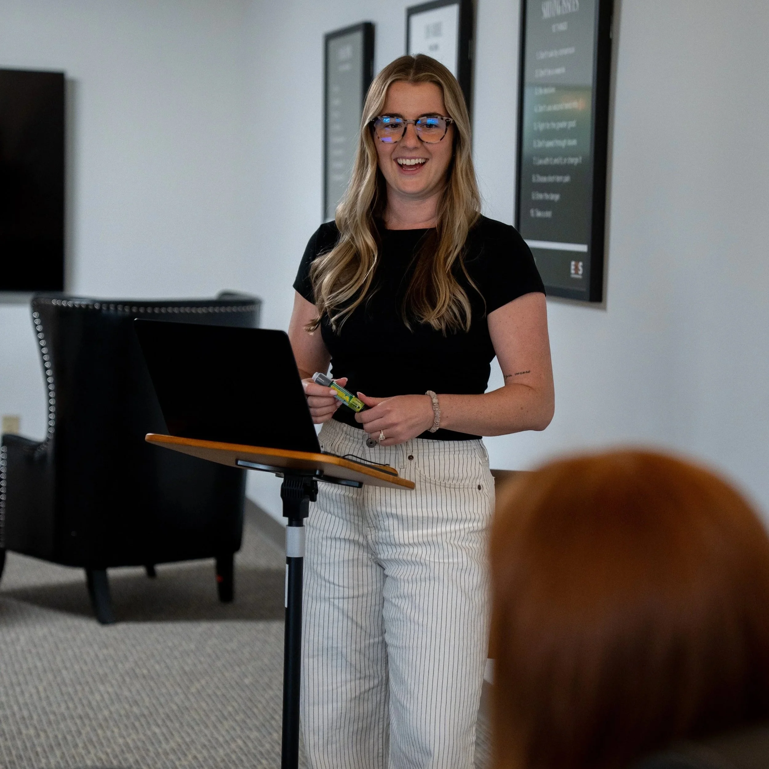A woman with long blonde hair, glasses, and a black shirt standing behind a podium or lectern in a room with framed posters on the wall. She is smiling and holding a yellow highlighter.