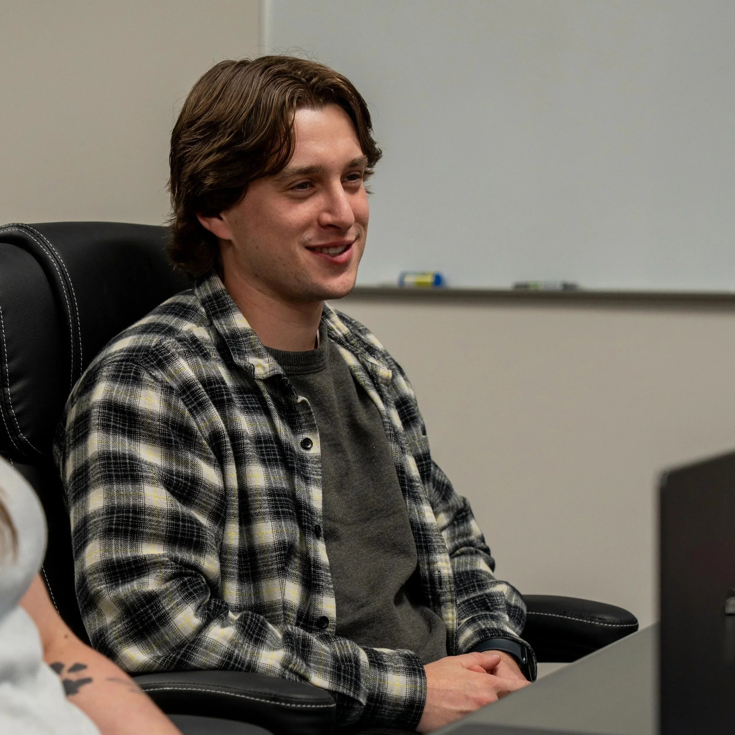 A young man with brown hair smiling, seated in a black office chair, in front of a plain light gray wall with a whiteboard and markers in the background, wearing a plaid shirt and a dark gray t-shirt.