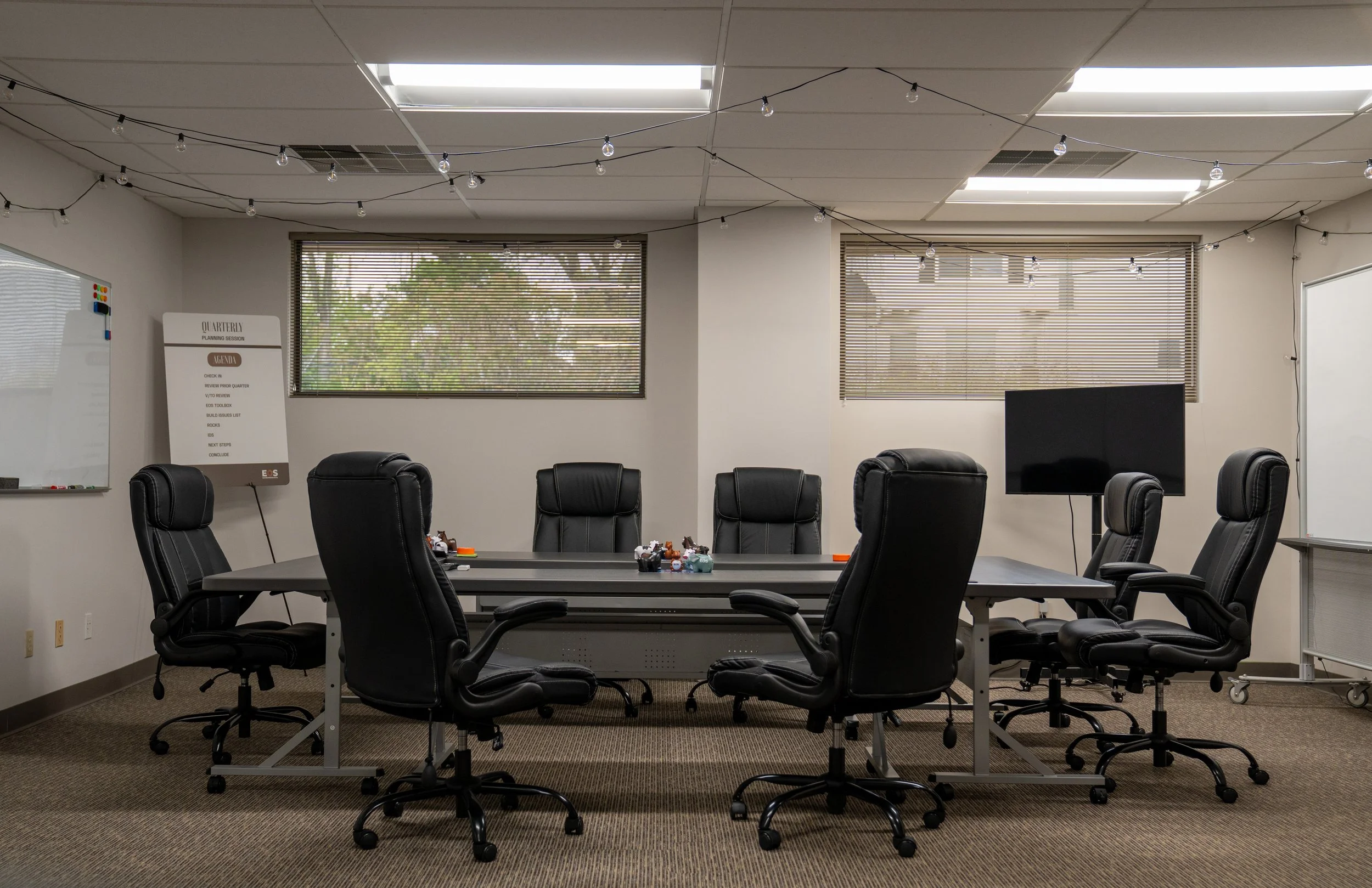 Conference room with a long table surrounded by black rolling chairs, two large windows with blinds, string lights hanging from the ceiling, a whiteboard, a large TV, and a whiteboard with an agenda.