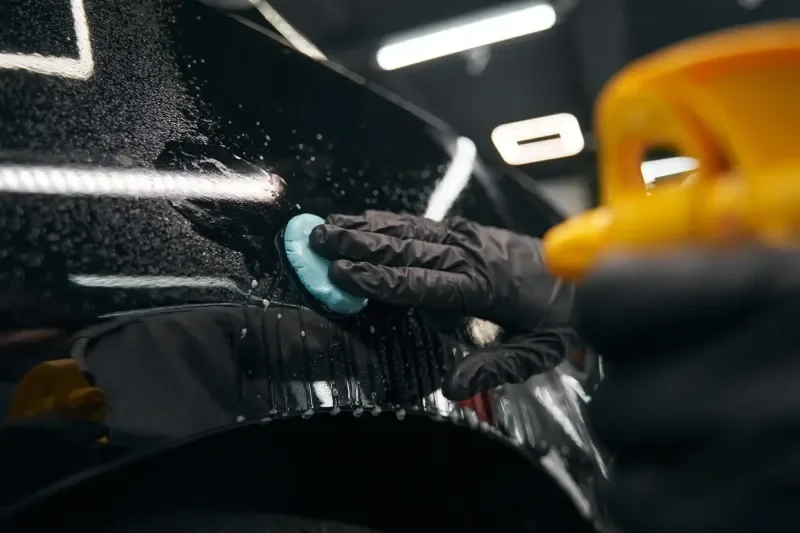 A person wearing black gloves polishing a black car with a clay bar in a garage or workshop.