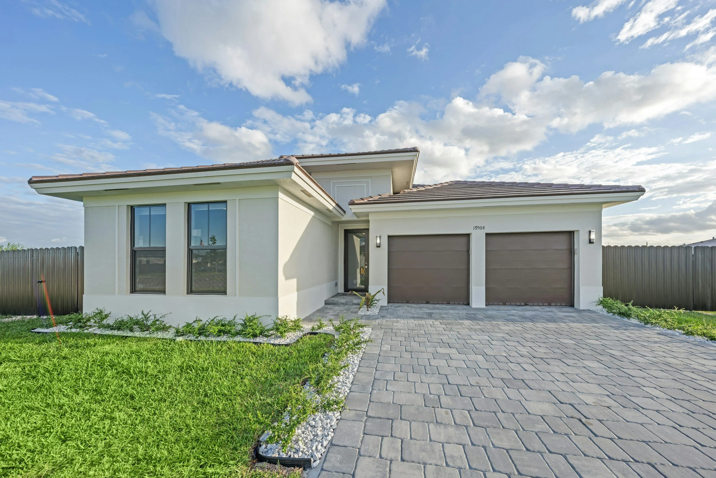Front view of a modern house with a double garage, light-colored exterior walls, two large windows, a paved driveway, and a small garden in the front yard, under a partly cloudy sky.