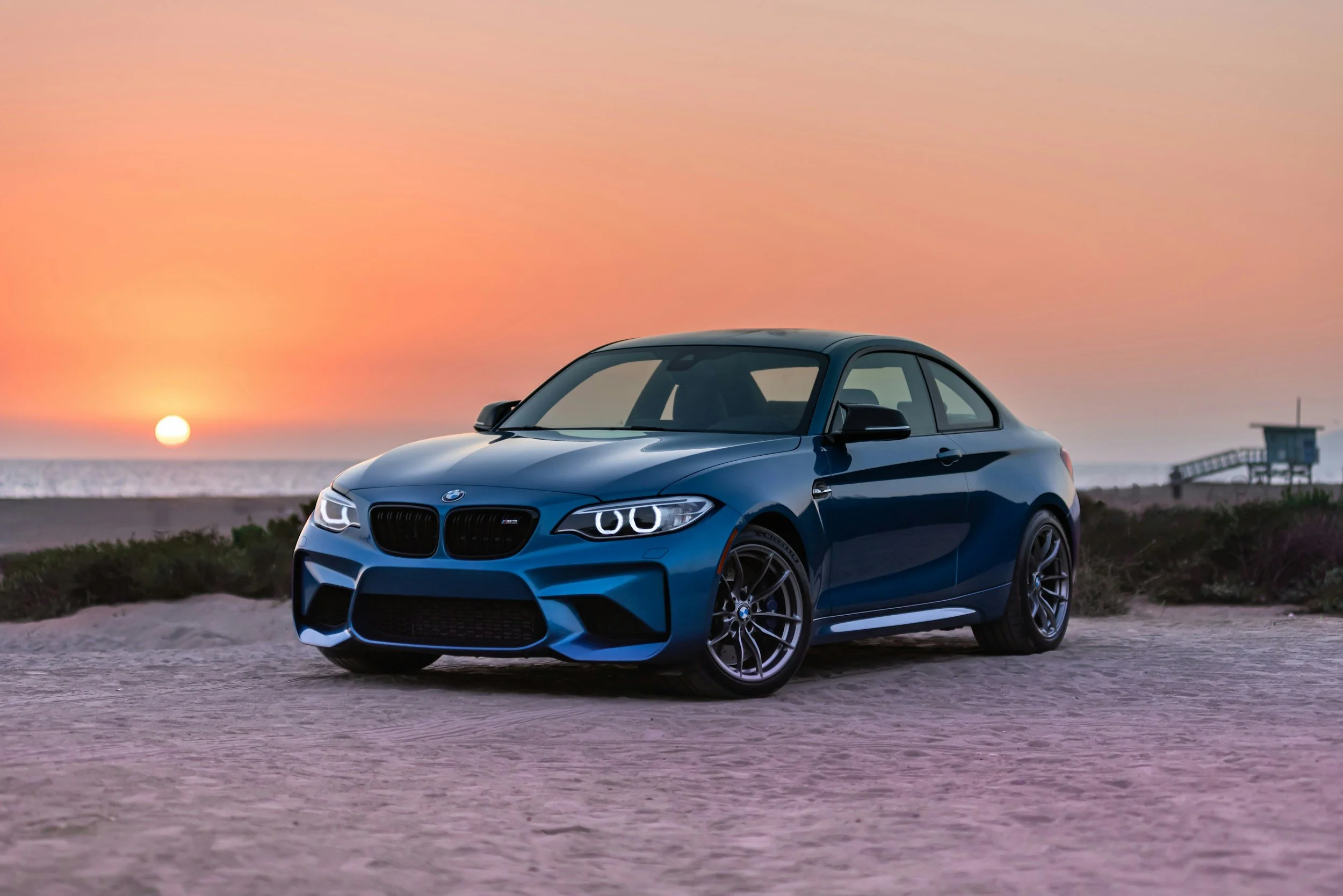 Blue BMW M2 sports car parked on a sandy beach during sunset with ocean and lifeguard tower in the background.