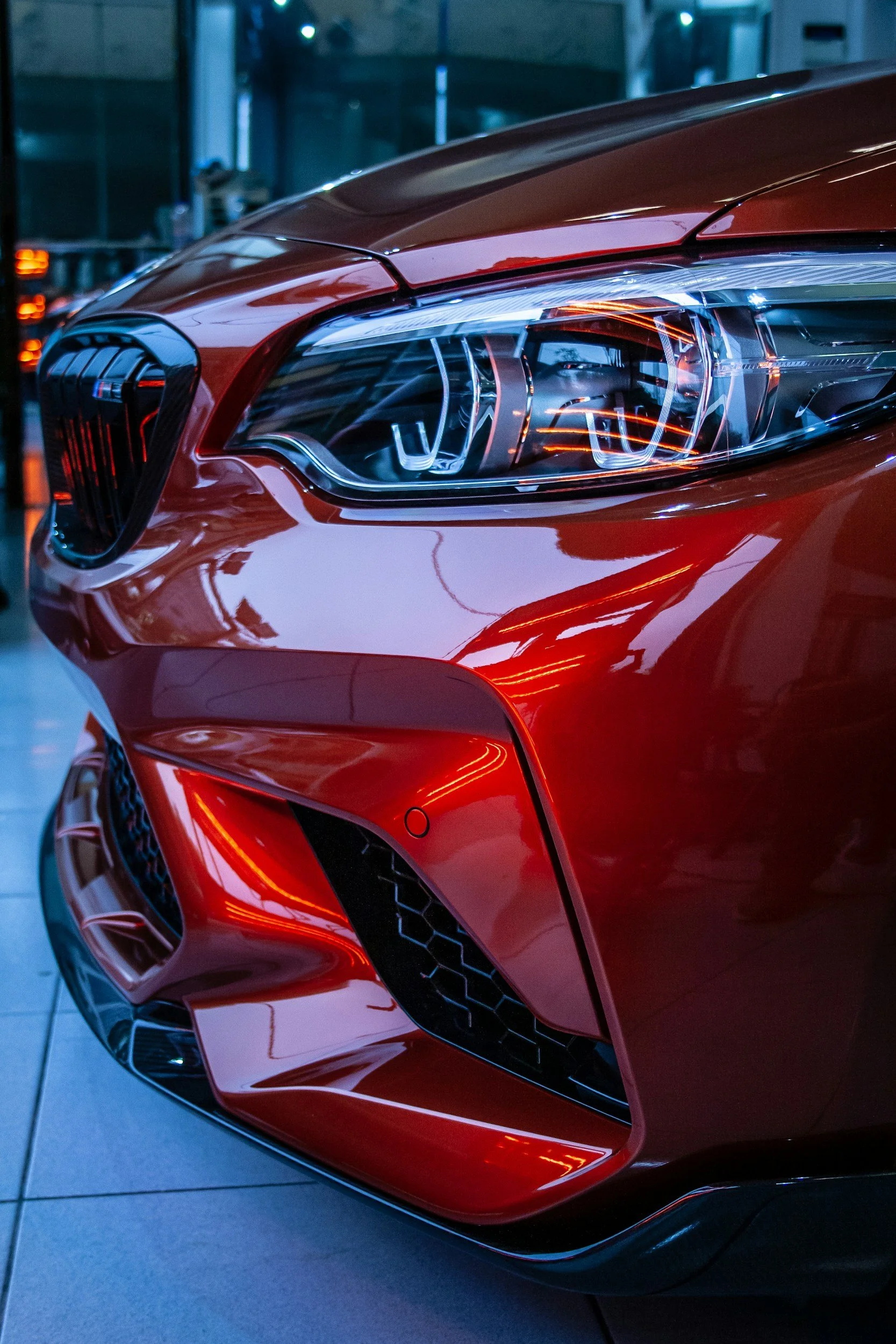 Close-up of a red luxury sports car's front, showing the headlight, grille, and bumper with detailed reflections and a modern design.