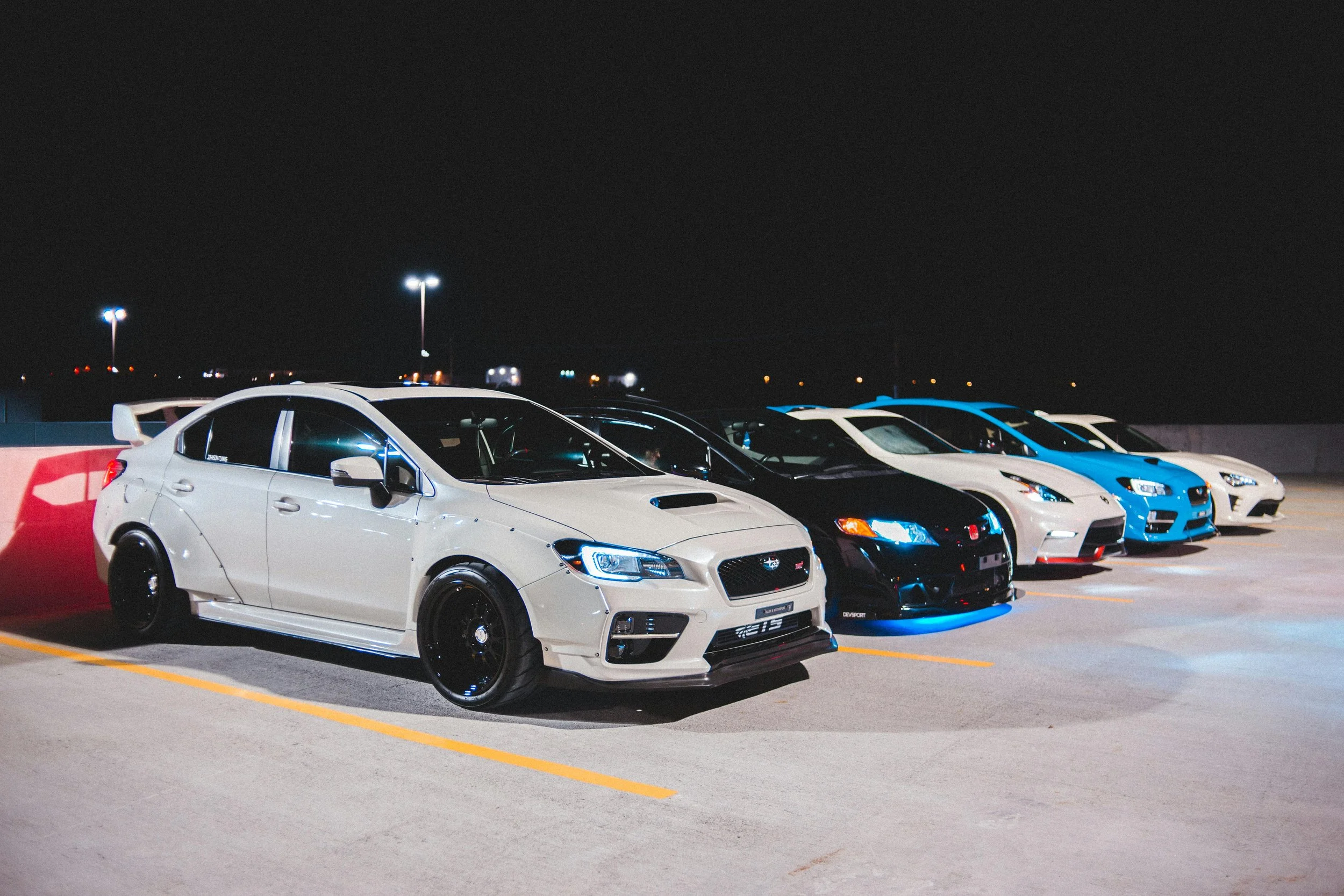 Lineup of six sports cars parked at night in a parking lot, illuminated by nearby lights, with a dark sky in the background.