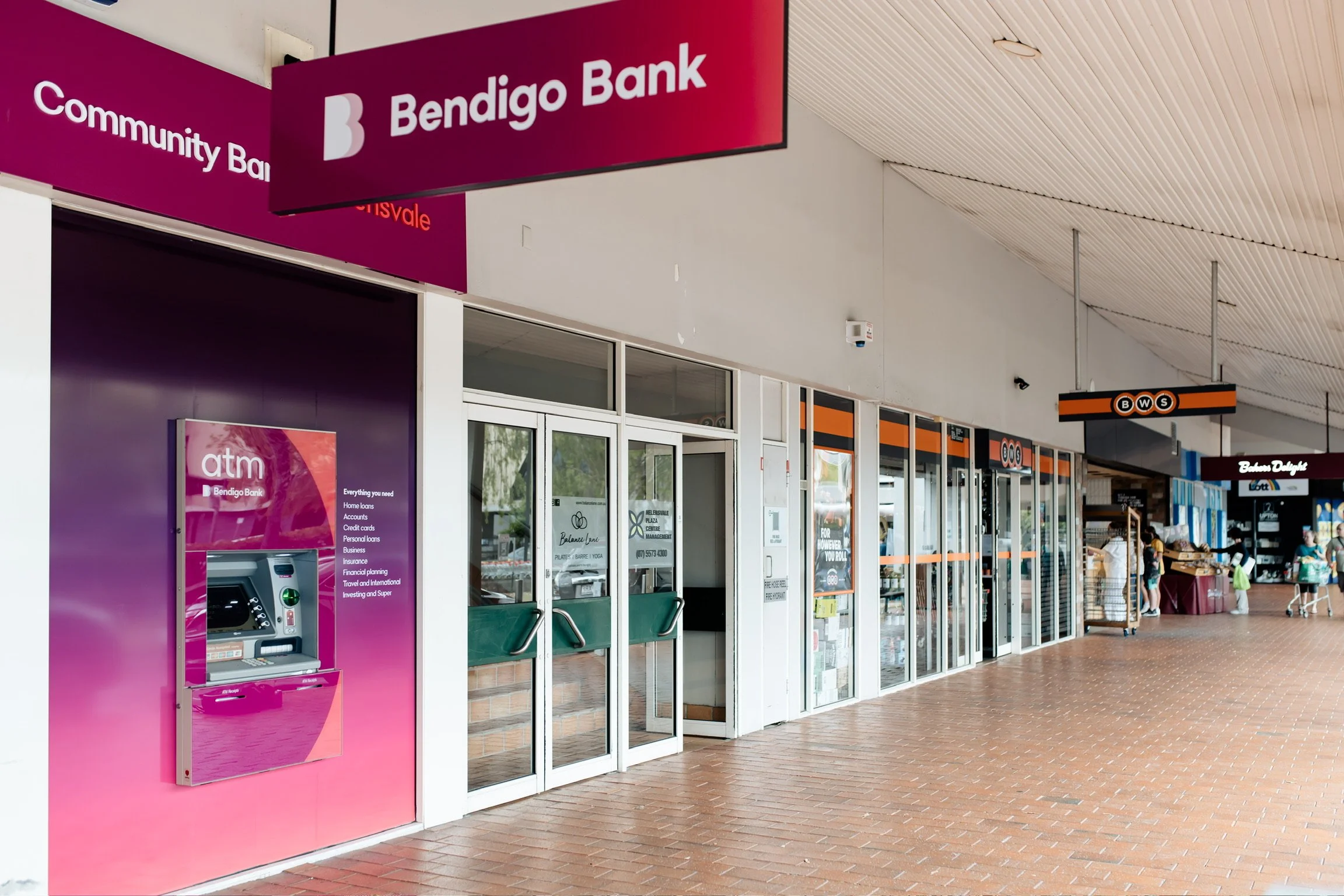 Exterior view of a shopping center with a Bendigo Bank branch, ATM machine, and People walking near shops and stalls.