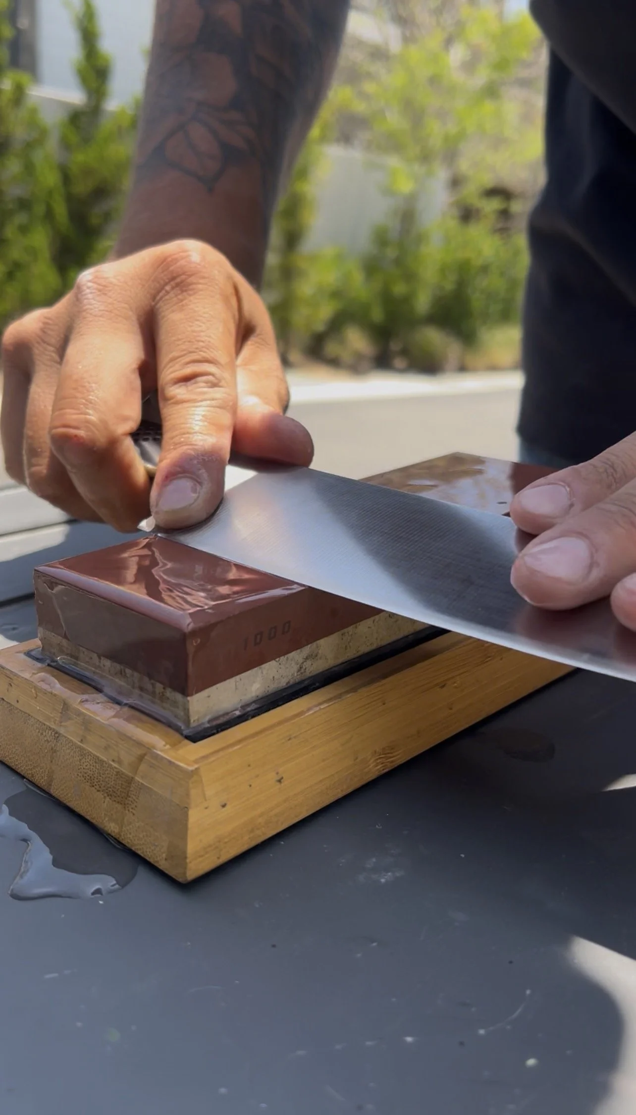 Close-up of a person's hand sharpening a knife on a sharpening stone outdoors on a wooden base, with a green natural background and sunlight. Knife sharpening in Newport Beach