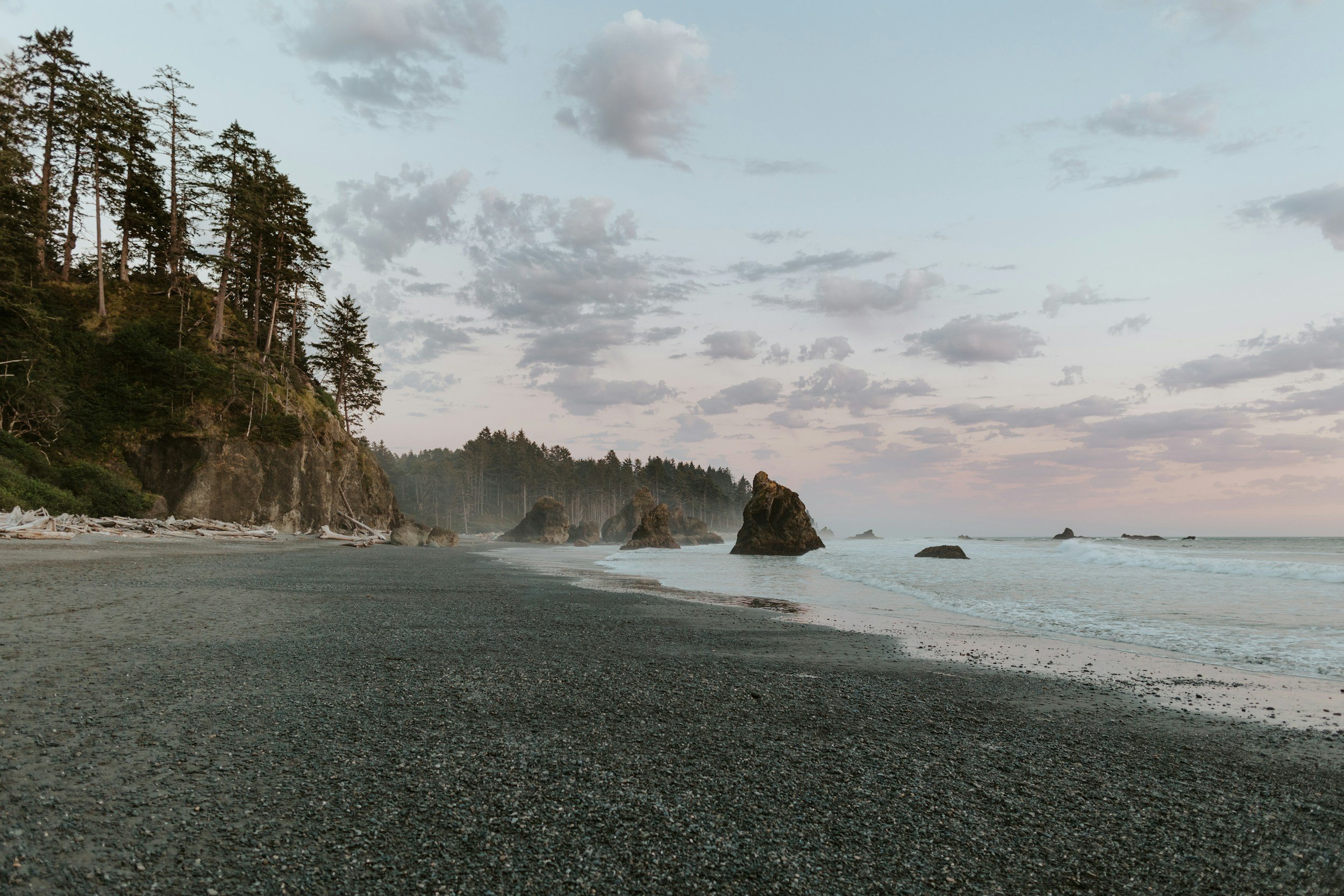 A secluded beach with dark sand, large rocks in the water, a forested cliff on the left, and a cloudy sky.