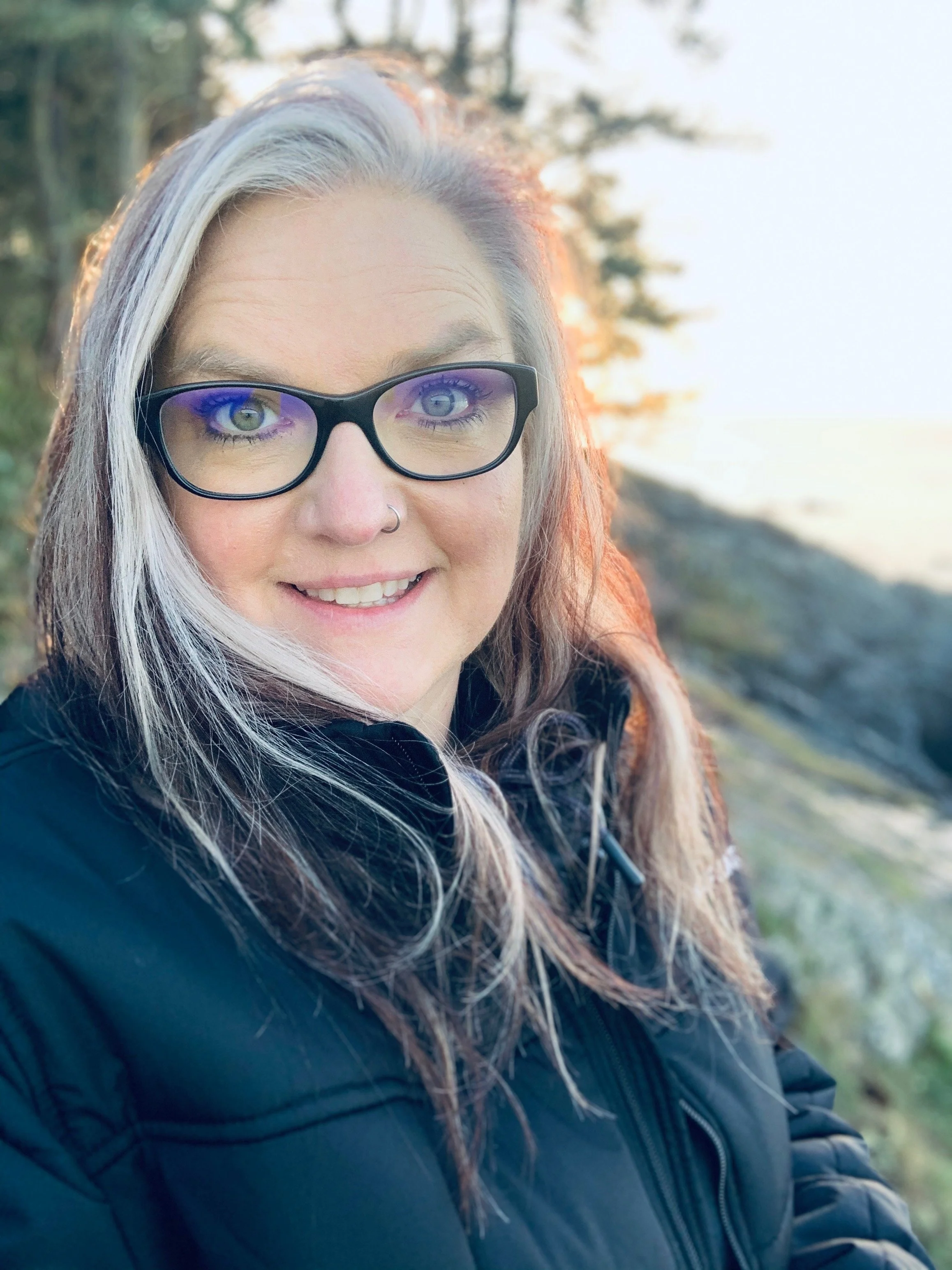 A woman with long brown and white hair, wearing glasses, smiling outdoors near a rocky shoreline with trees in the background at sunset.