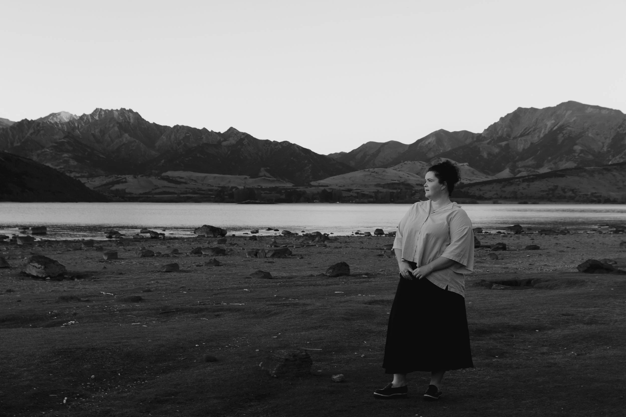 A woman standing on a rocky shoreline near a body of water with mountains in the background, in black and white.