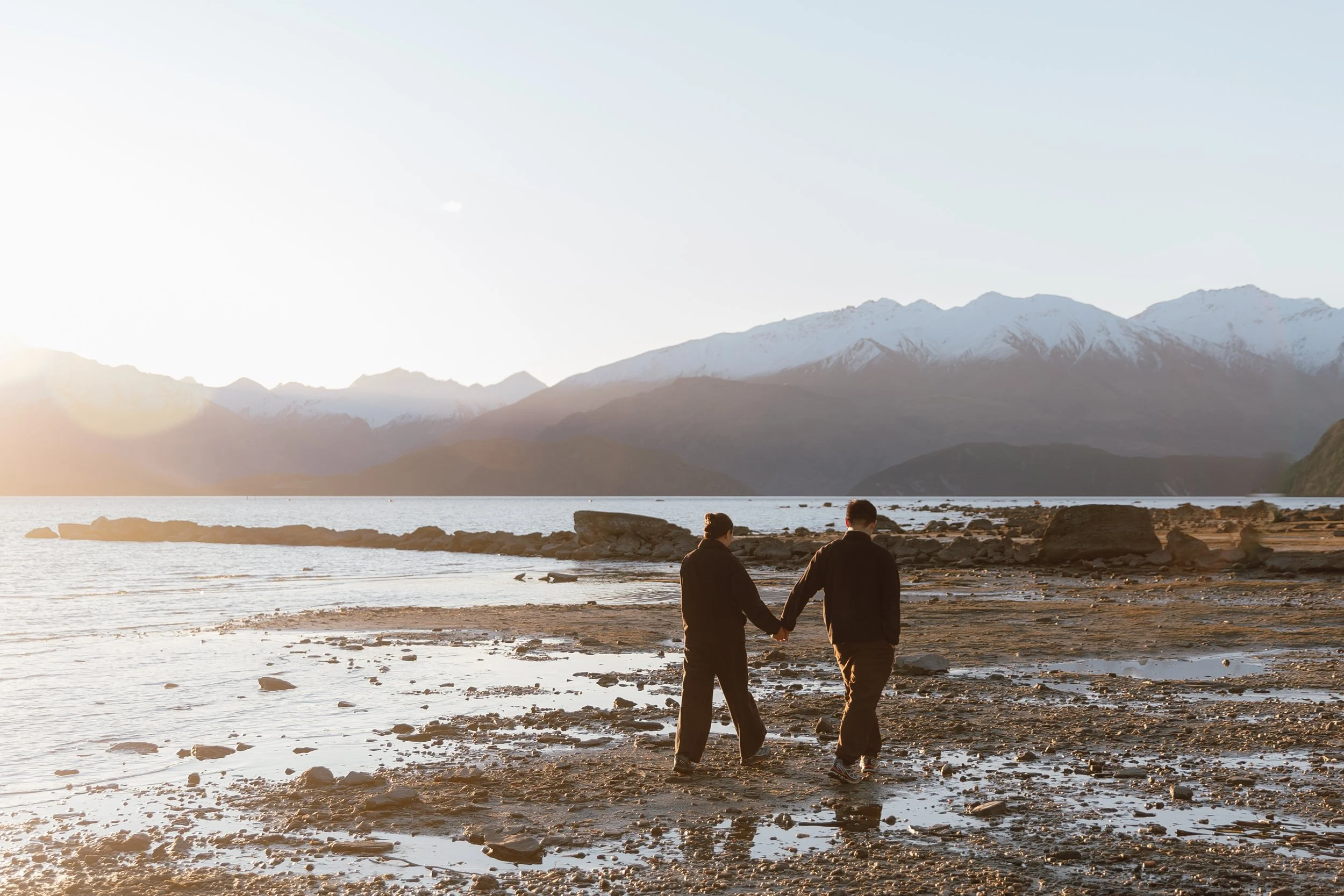 A couple holding hands walking along a rocky shoreline at sunset, with mountains and snow in the background.