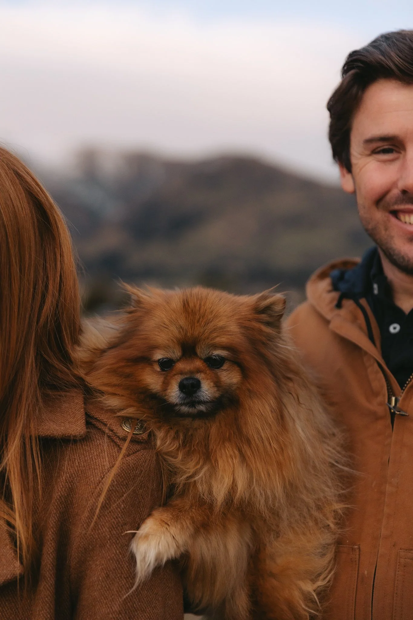 Close-up of a fluffy brown dog with black eyes being held by a person with long red hair, wearing a brown jacket. Part of a smiling person's face is visible on the right. The background is outdoor with blurred scenery.