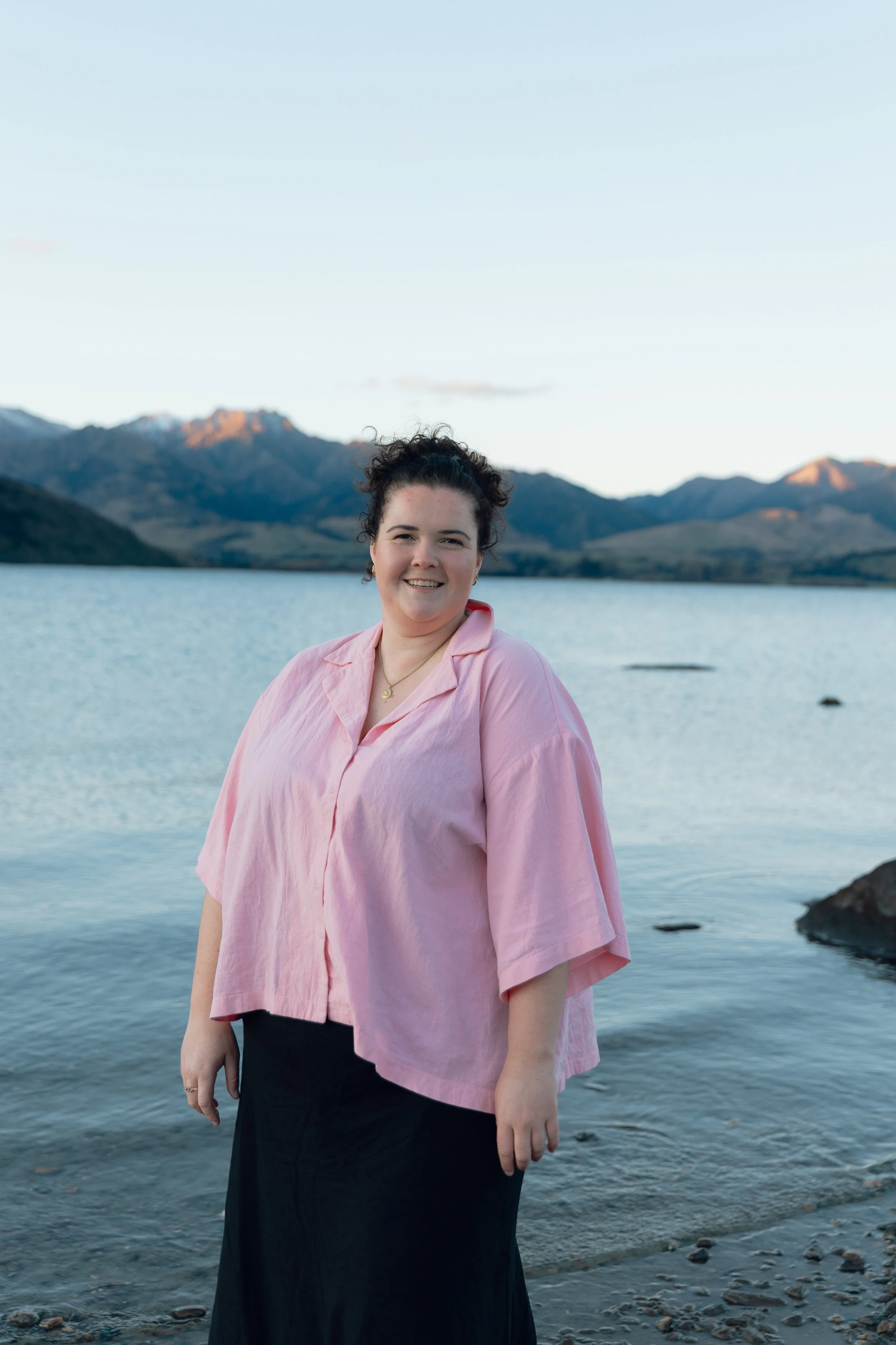 A woman with dark curly hair, wearing a pink blouse and a black skirt, standing by a lake with mountains in the background during dusk.