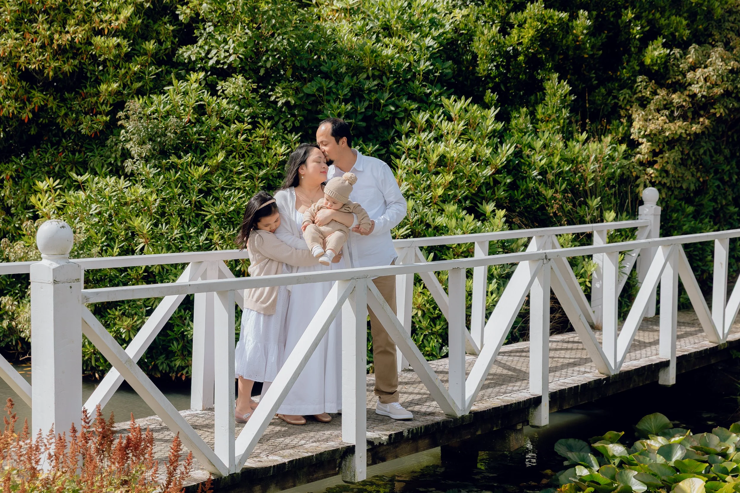 A family of four standing on a white bridge over water with green bushes and lily pads in the background. The parents are holding a baby, and their daughter is hugging her mother.