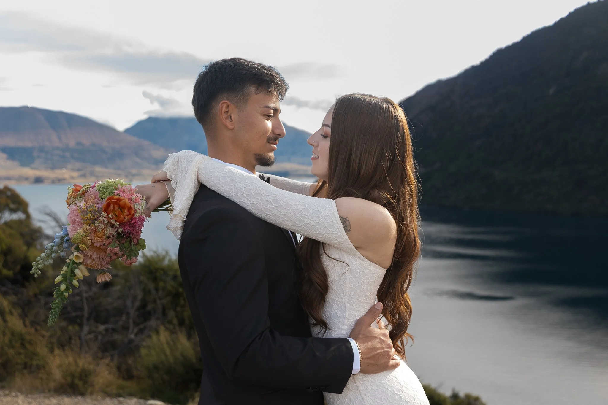 A newlywed couple embracing outdoors with mountains and water in the background. The groom is wearing a black suit and holds a bouquet of pink, orange, and green flowers. The bride wears a white lace wedding dress, and both are smiling.