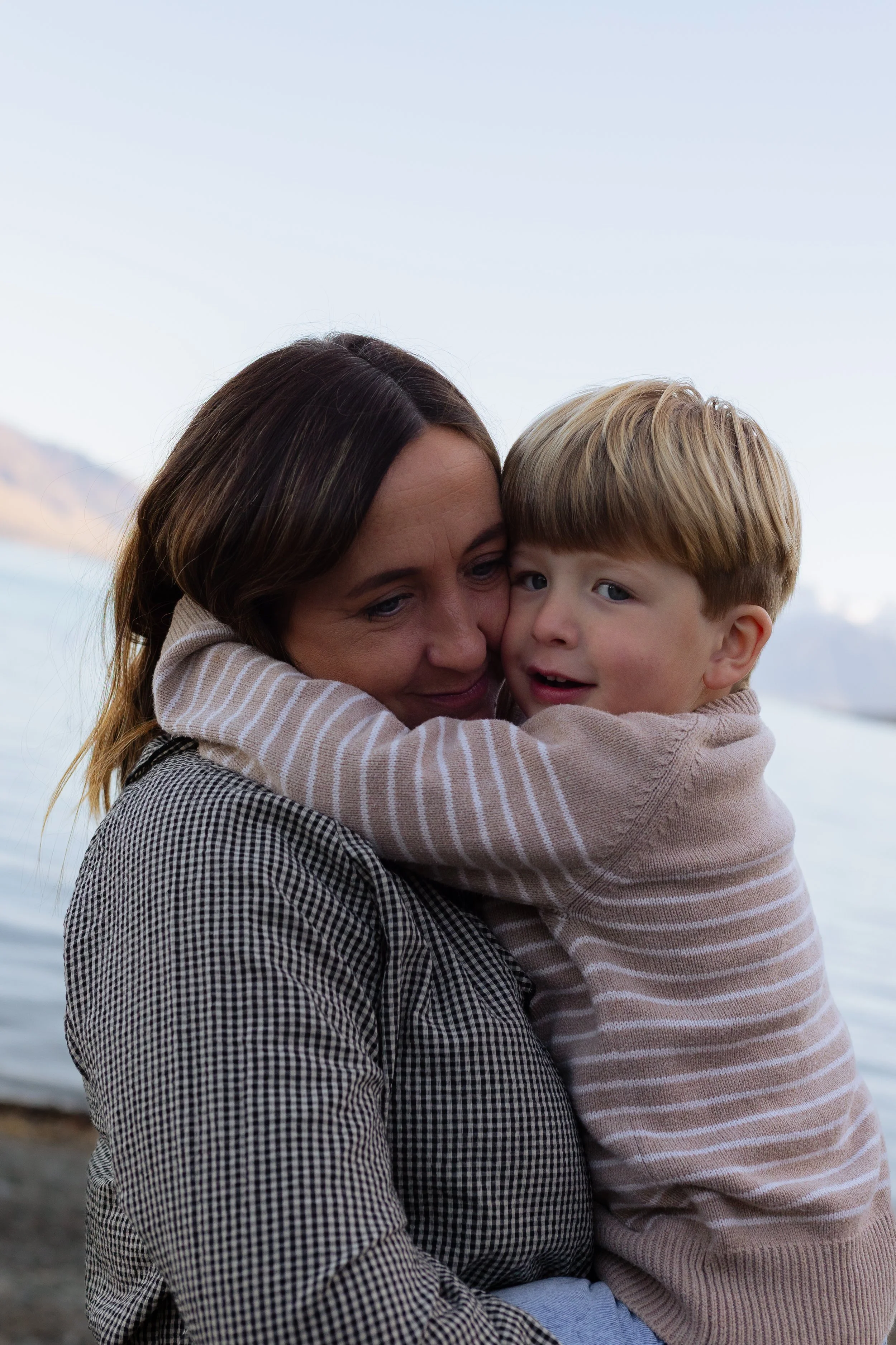 A woman and a young boy hugging outdoors near a body of water with mountains in the background.