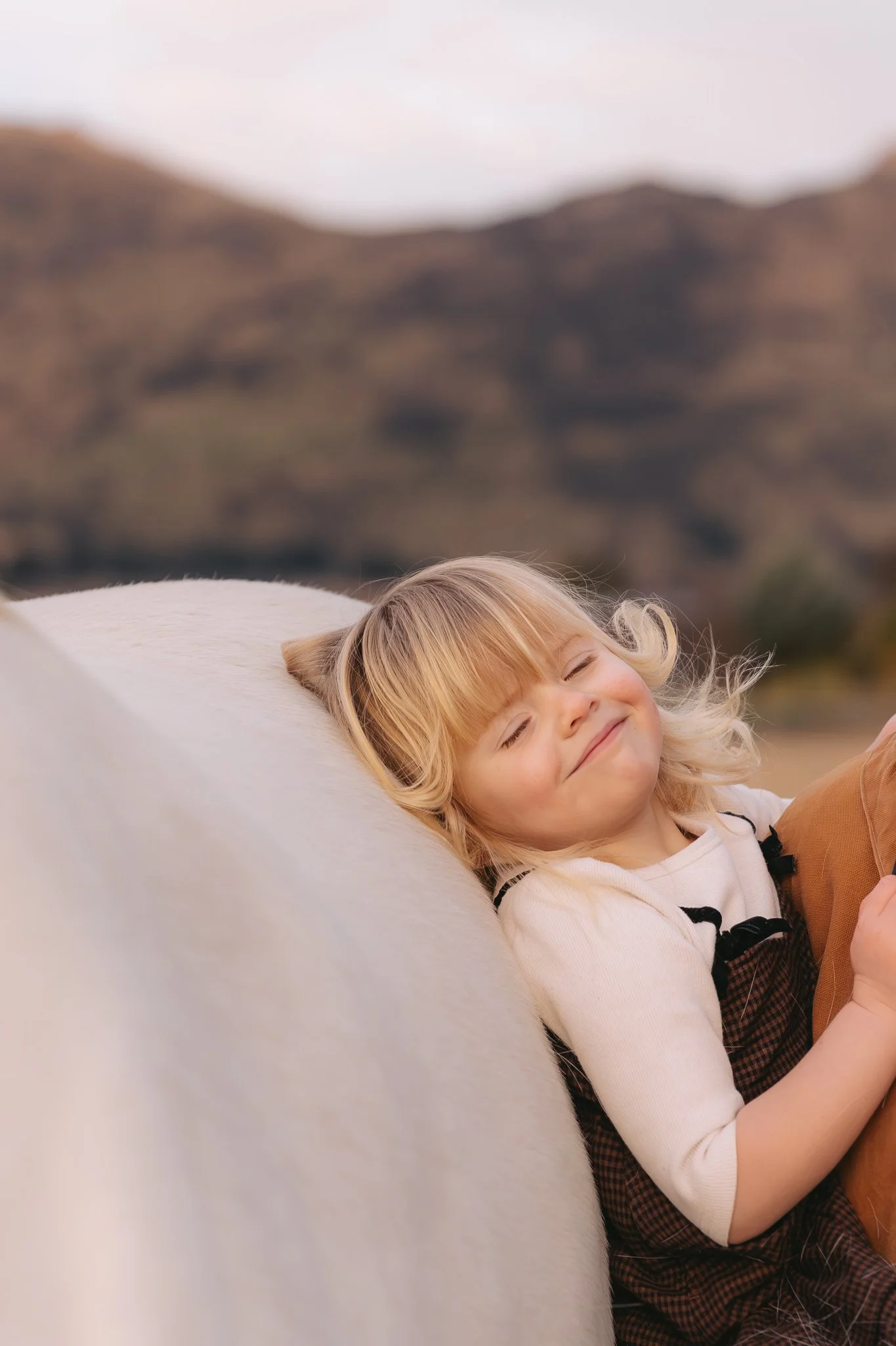 A young girl with blonde hair and a big smile is lying down on a white horse's back, outdoors with hills in the background.