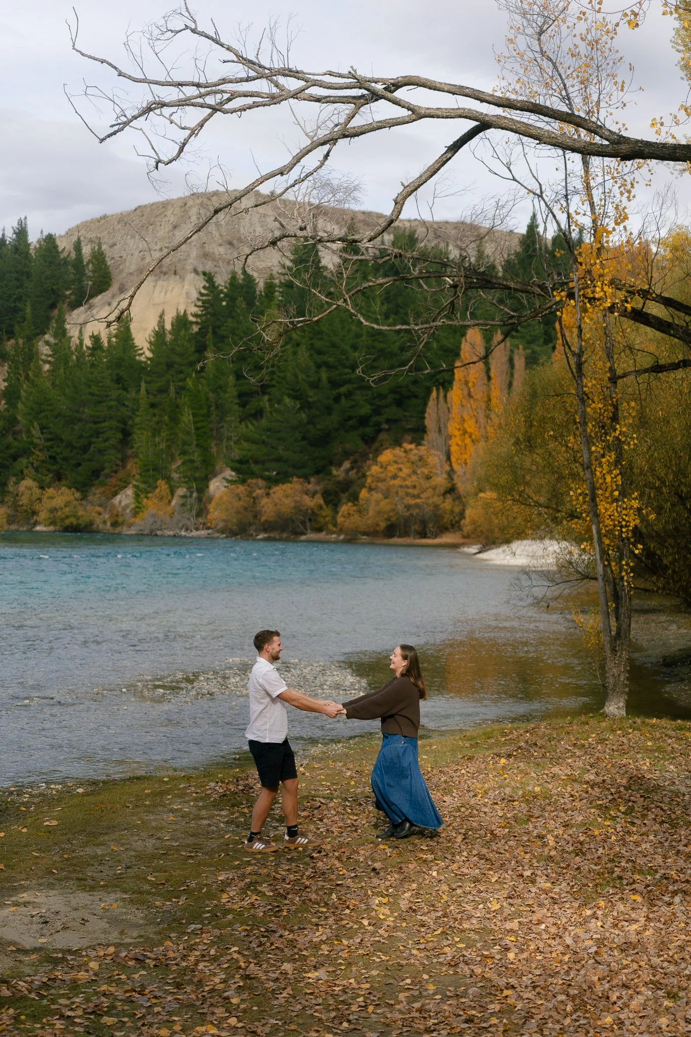 A man and a woman holding hands by a river, surrounded by fall trees and mountains, with a branch overhead.