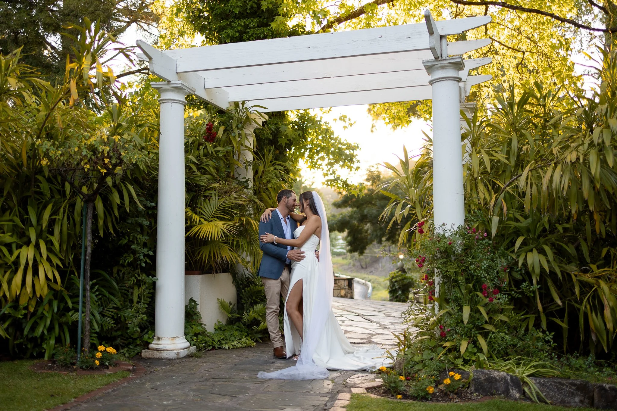 Bride and groom embracing in wooden arch. evening light making vibrant green garden