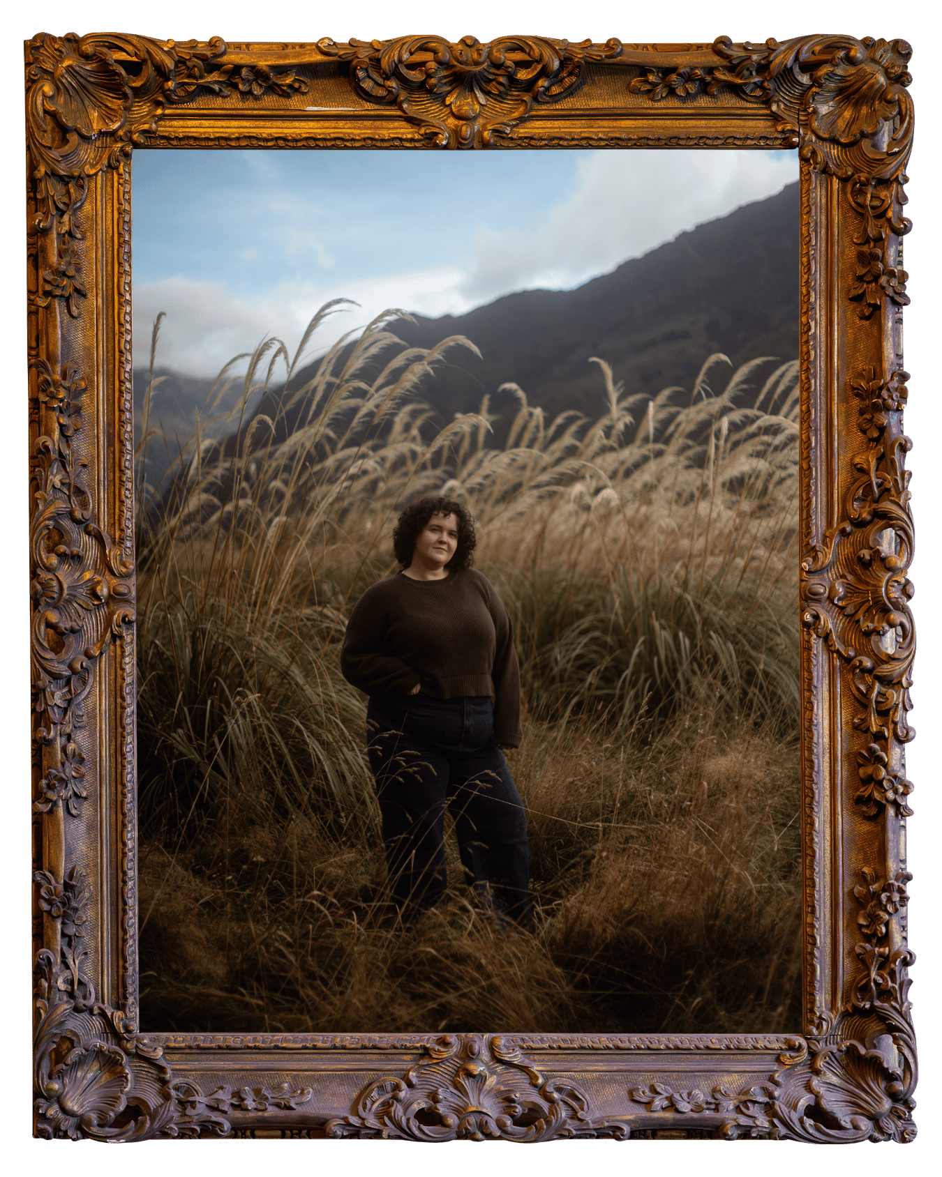 A woman standing in a grassy field with a mountain range in the background, seen through an ornately carved wooden picture frame.