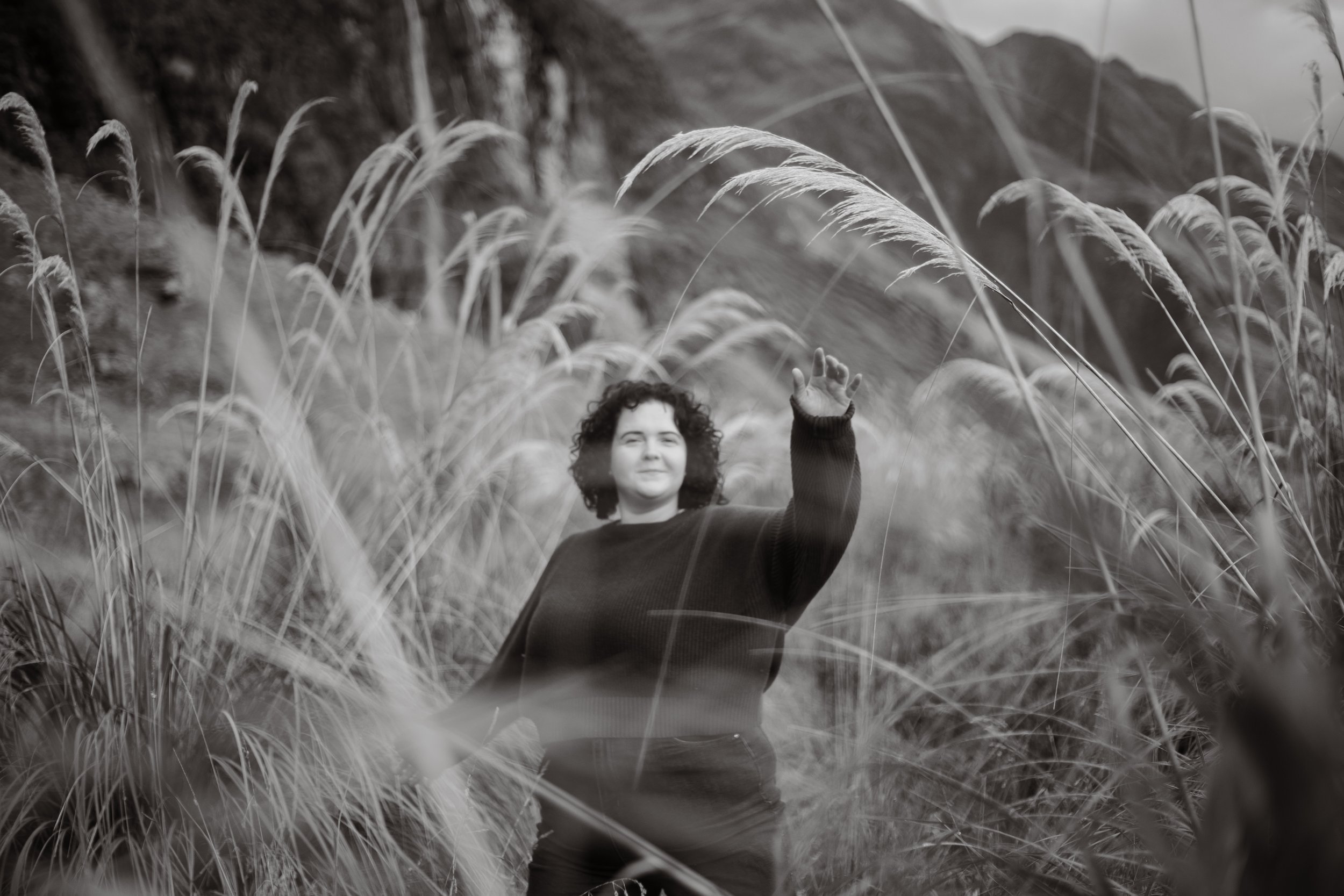 Black and white image of girl below mountains in wanaka, surrounded by long grass and flaxes.