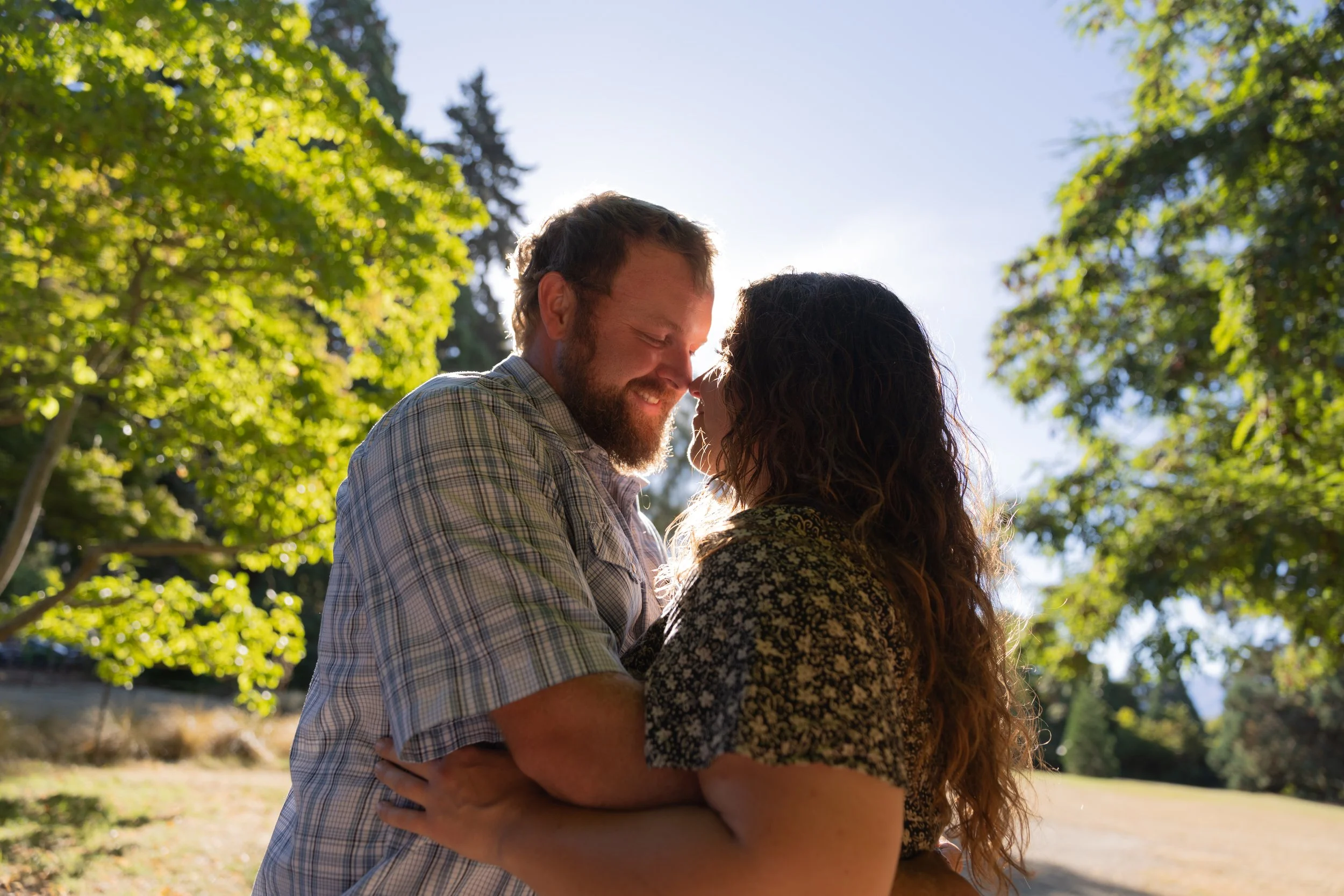 A couple in a park, smiling and touching foreheads, with sunlight filtering through trees in the background.