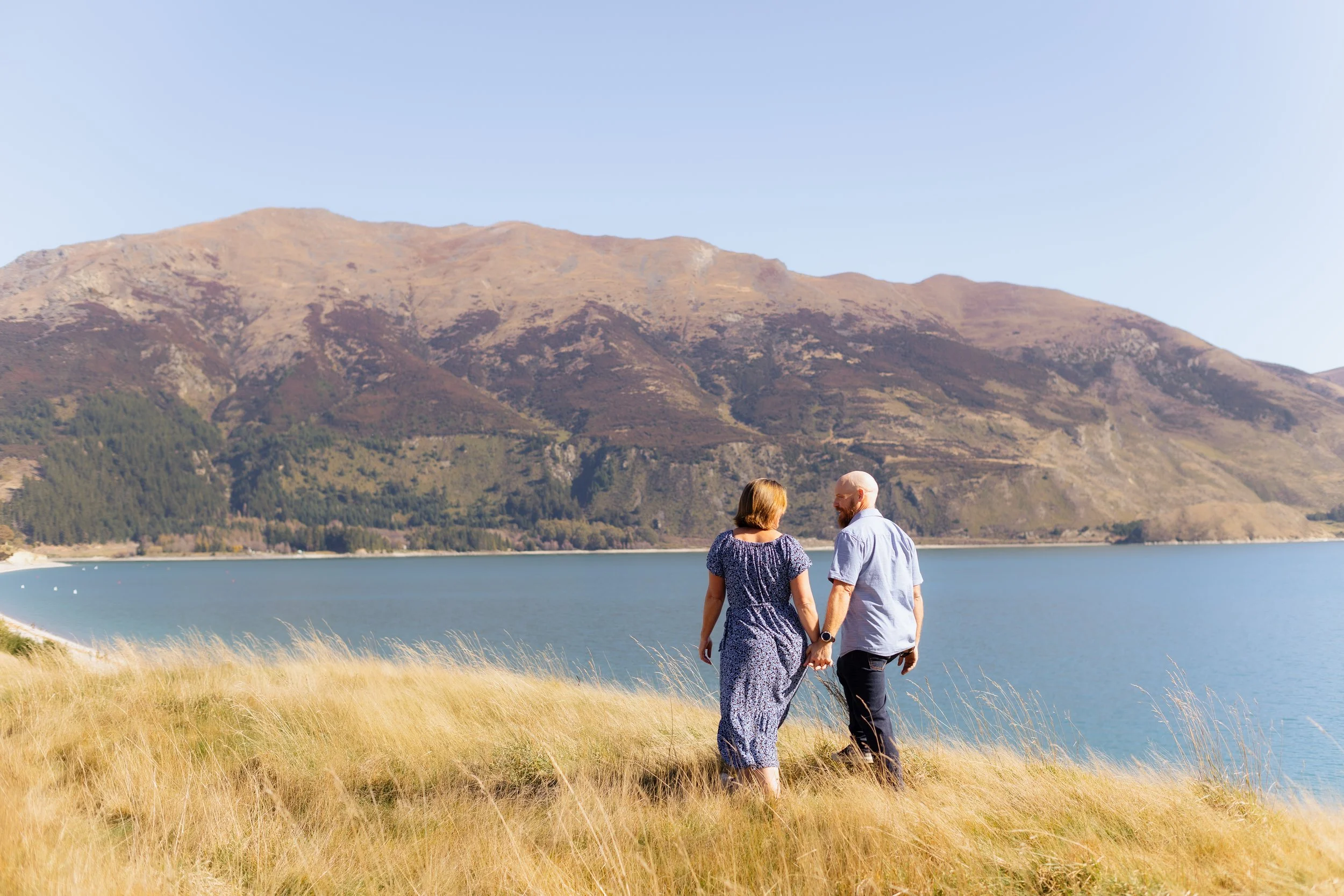 A couple walking hand in hand along a lakeshore with mountain in the background on a clear day.
