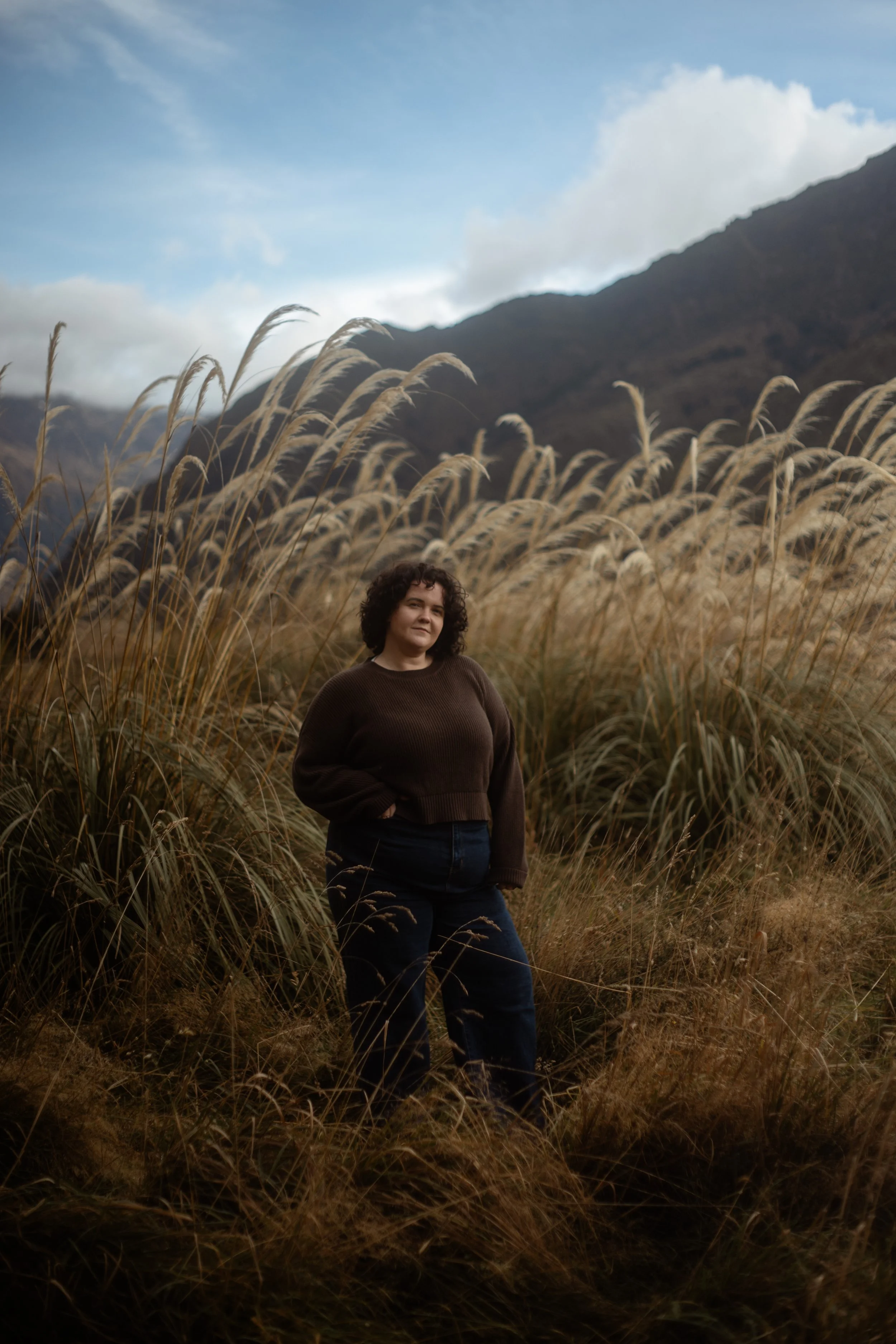 Woman with curly hair standing below wanaka mountains amongst long grass and toi toe's.