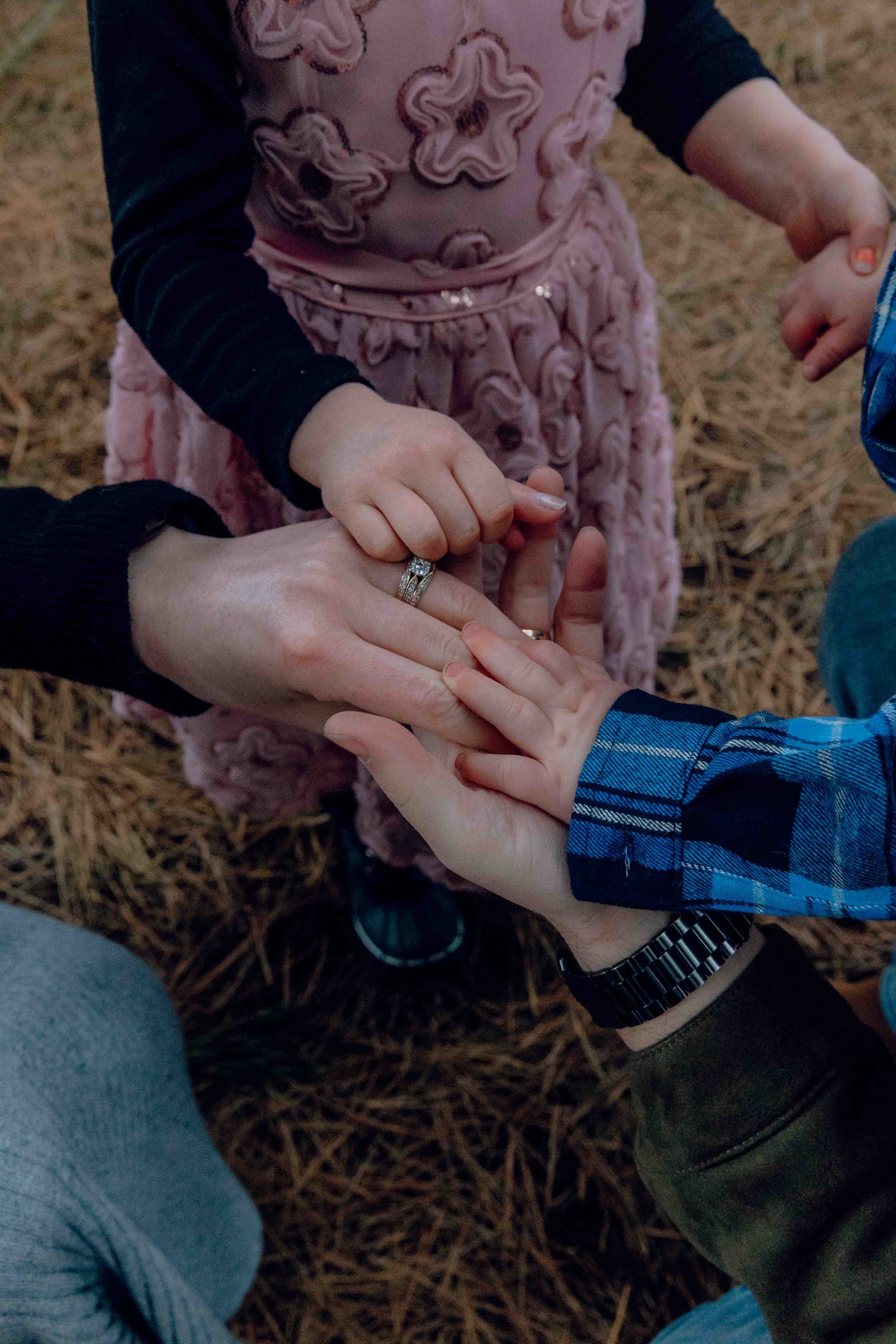 Multiple hands, including a child's, holding and touching a woman's hand with a wedding ring, on a background of pine needles.
