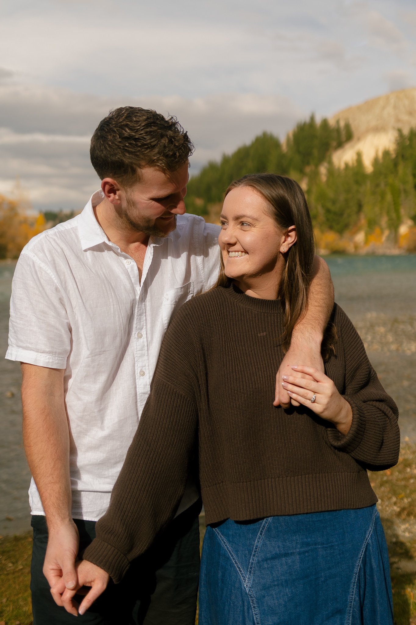 A smiling couple outdoors with a river, trees, and mountains in the background, embracing each other.