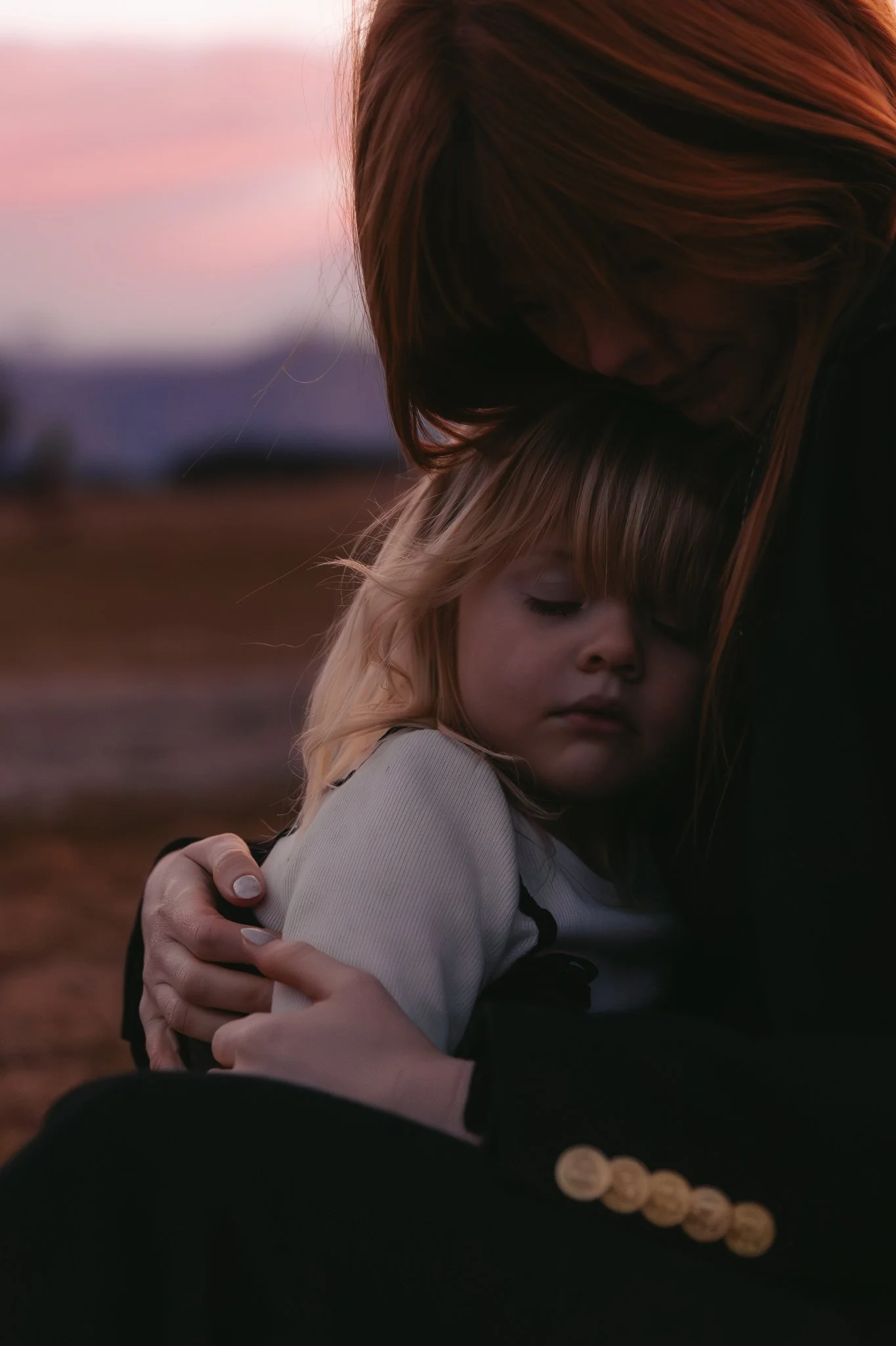A woman with long red hair hugging a young girl with blonde hair outdoors during sunset, with the woman's face tucked into the girl's hair, creating a tender moment.