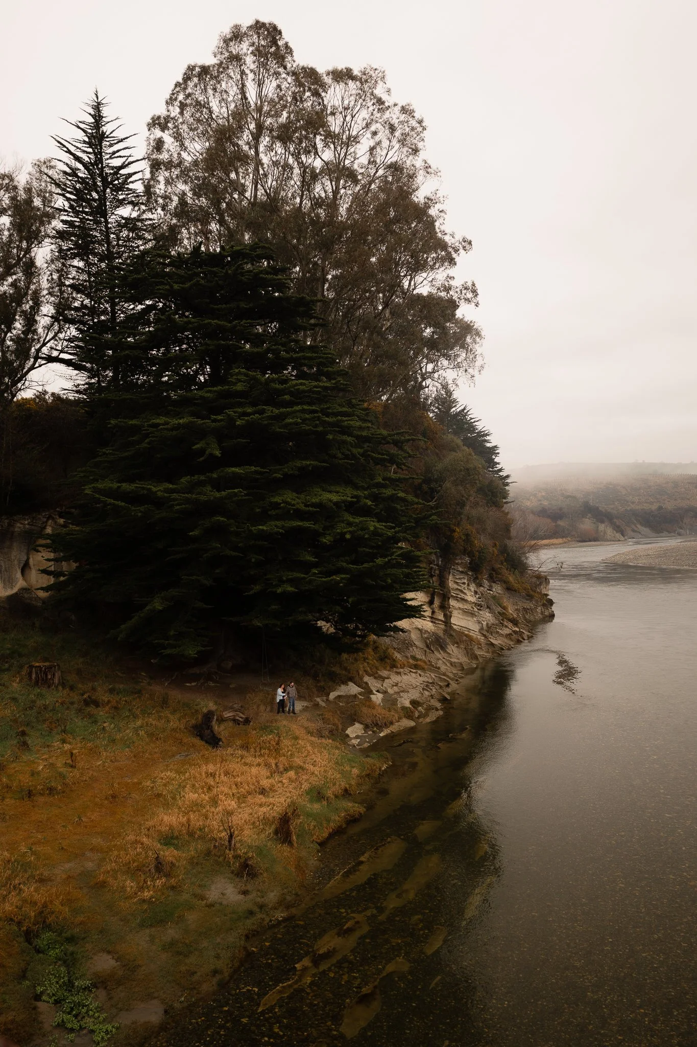A river bank with trees and a cloudy sky, two people standing near the water on rocks.