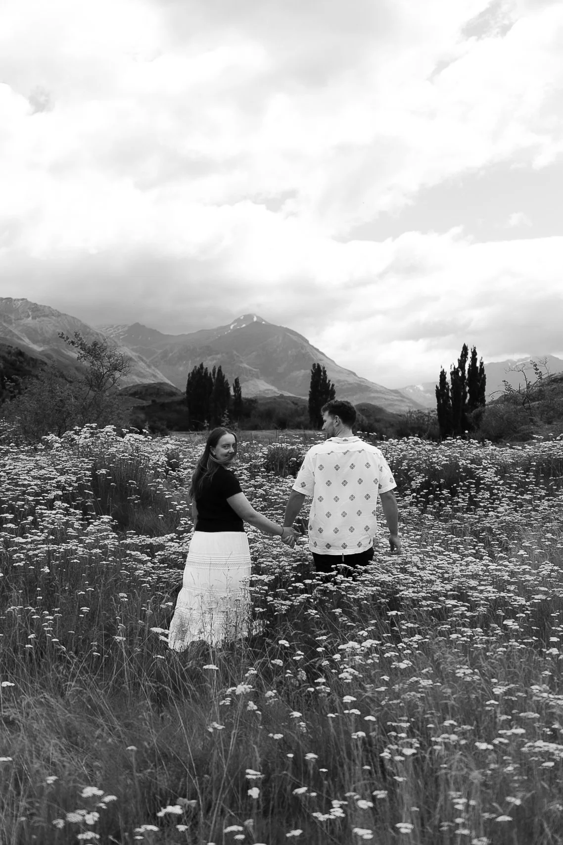 A black and white photo of a couple holding hands and walking through a field of flowers with mountains in the background.