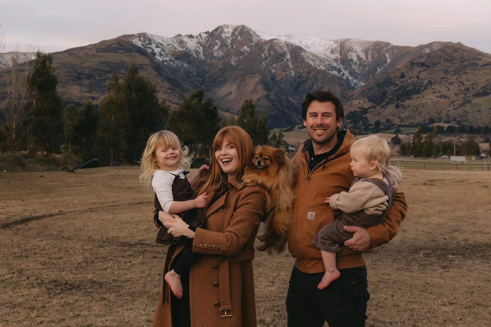 A family of four with a small dog smiling outdoors with a mountainous landscape in the background, during late afternoon or early evening.