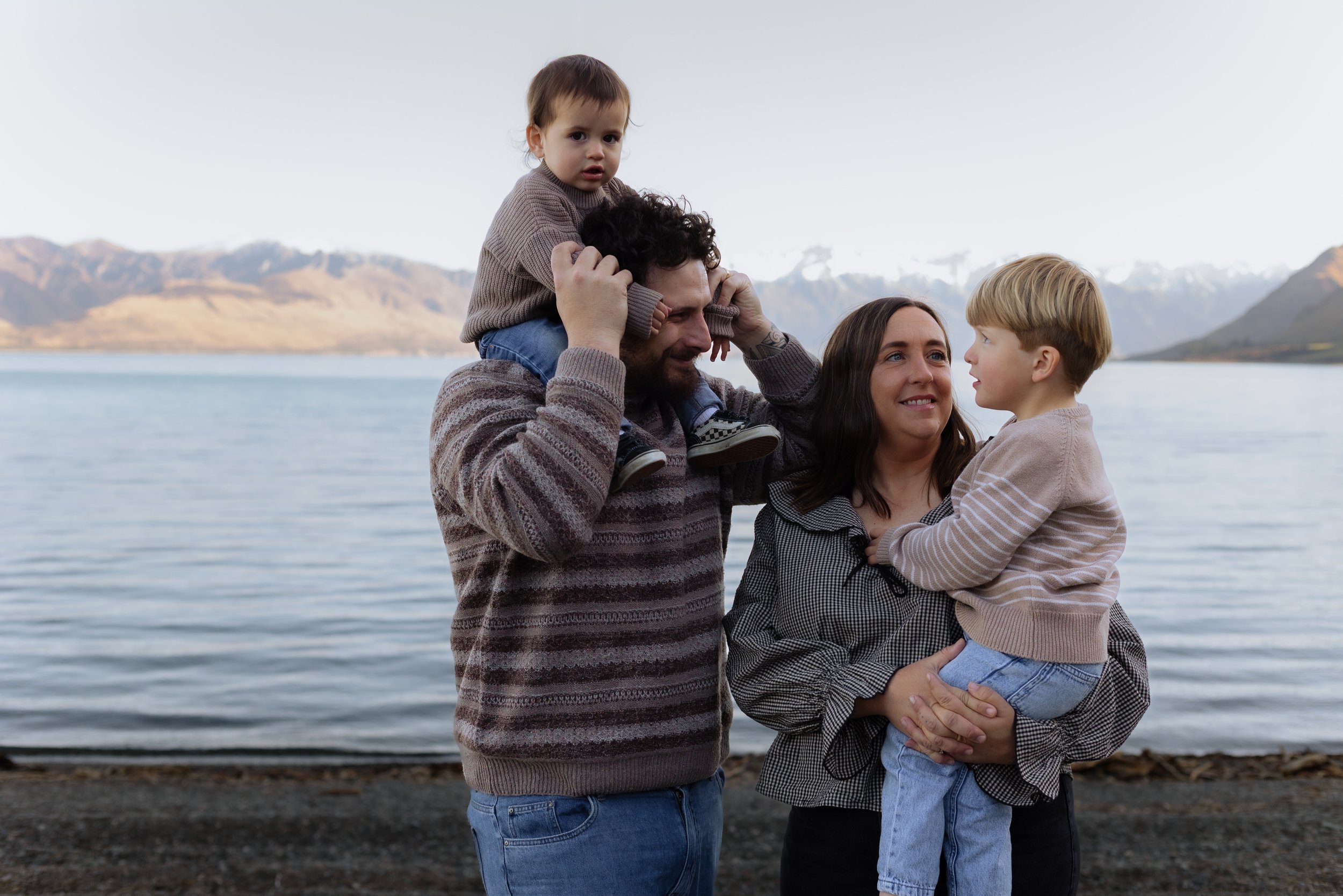 A family of four stands near a body of water with mountains in the background. The father, with curly dark hair and beard, is carrying a young girl on his shoulders. The girl, with short dark hair, is wearing a brown sweater. The mother, with long da