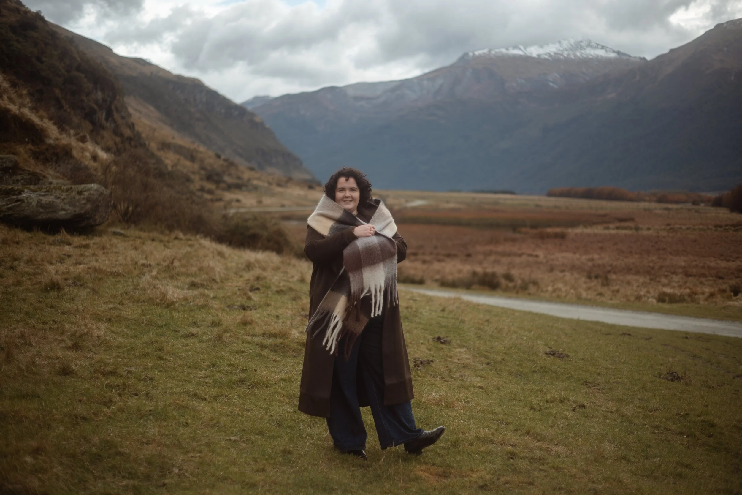 curly haired female dressed in brown trench coat and warm scarf standing surrounded by mountains
