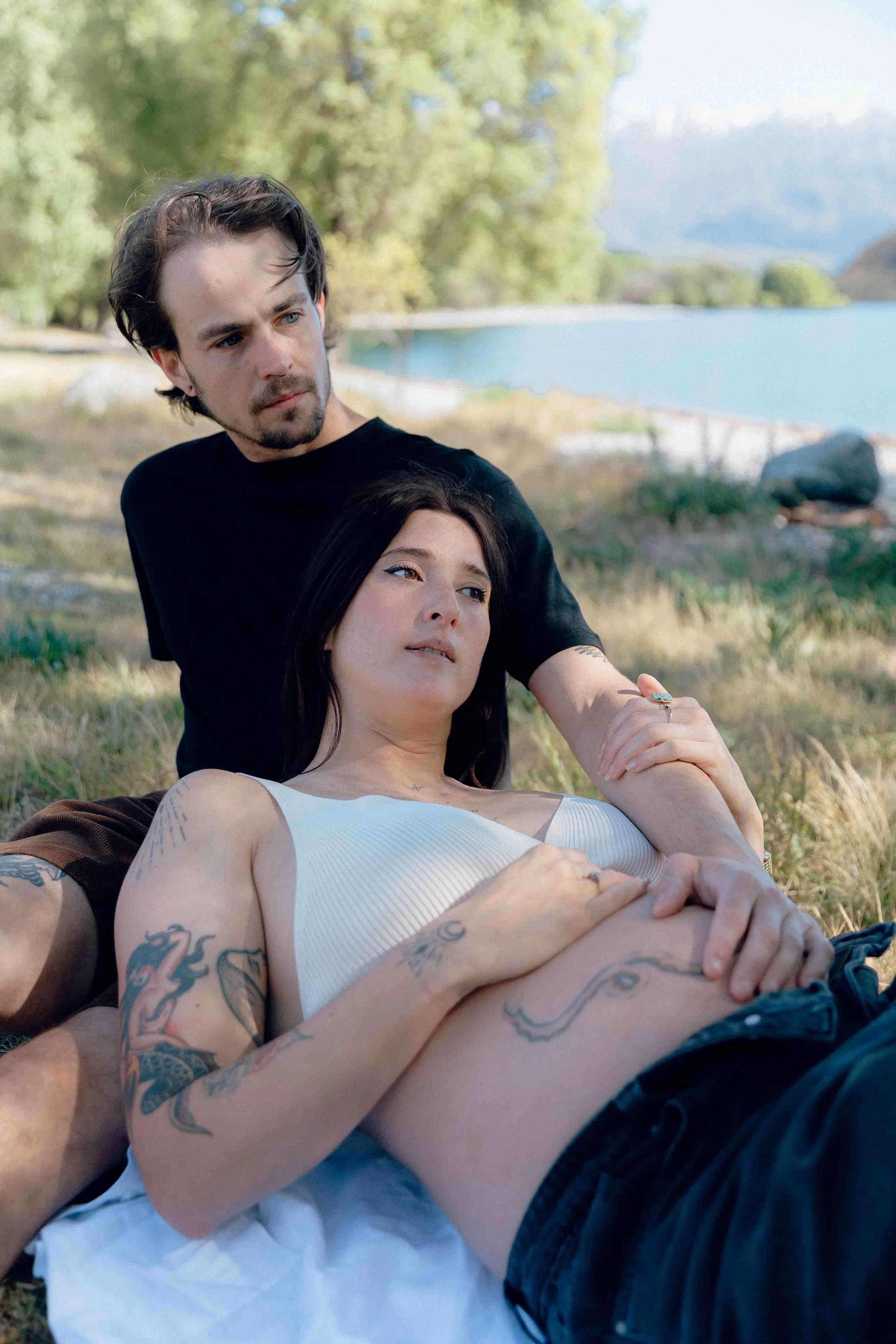 A young man and woman lying on the grass near a lake with trees and rocks in the background. The woman has tattoos and is wearing a white top, while the man is in a black t-shirt.