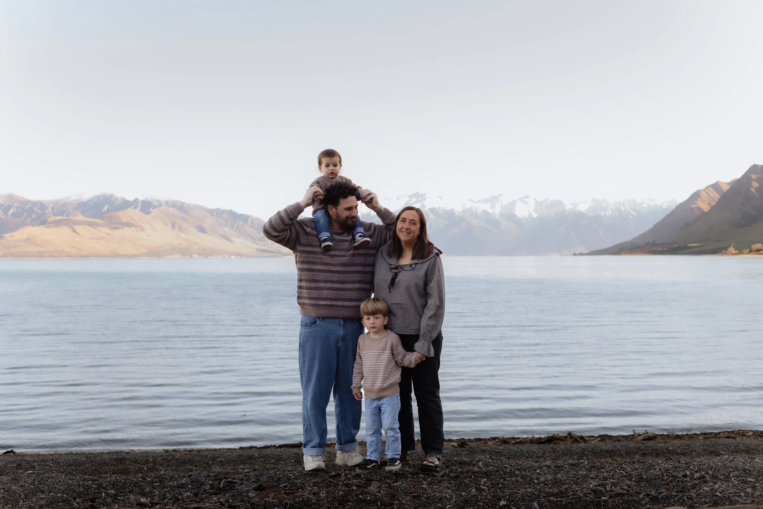 A family of four standing on a rocky shoreline near a calm lake with mountains in the background. The man is holding a child on his shoulders, and a woman and another child are standing beside him, all smiling and wearing casual clothing.