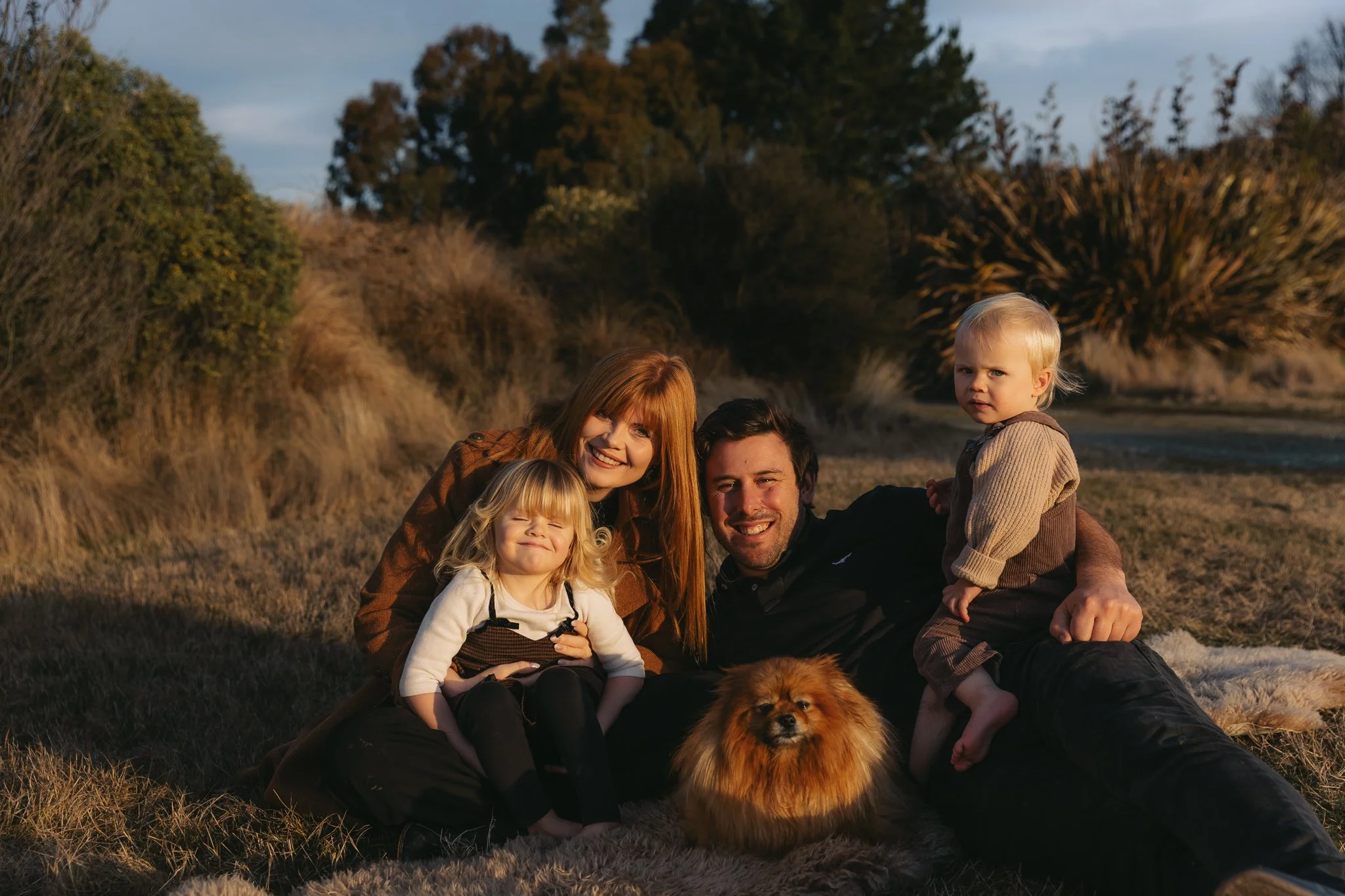 A family of five and a dog sitting outdoors on grass in a natural setting during late afternoon with trees and a cloudy sky in the background.