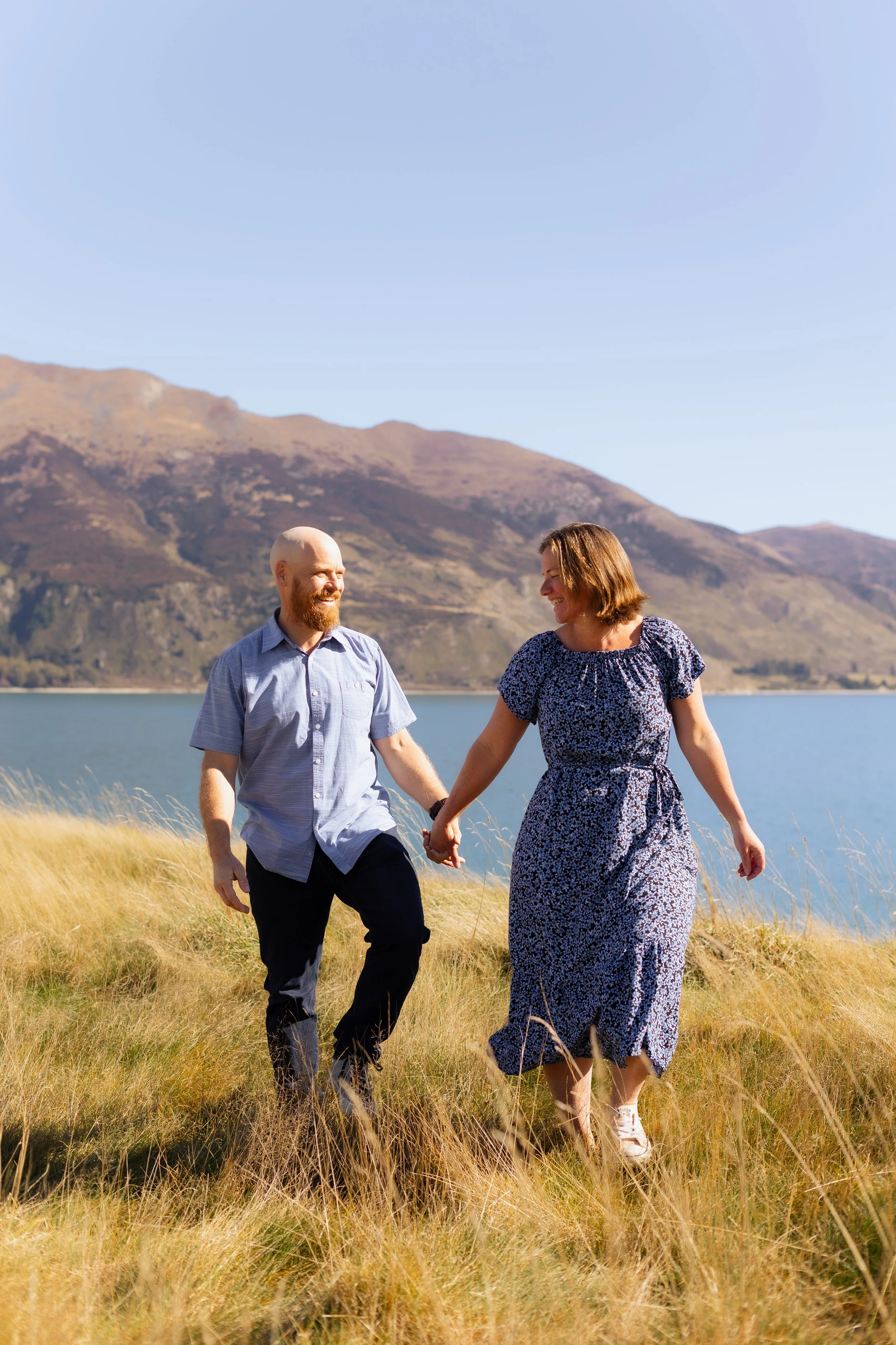 A couple holding hands and walking through a field of tall grass near a lake with mountains in the background under a clear sky.