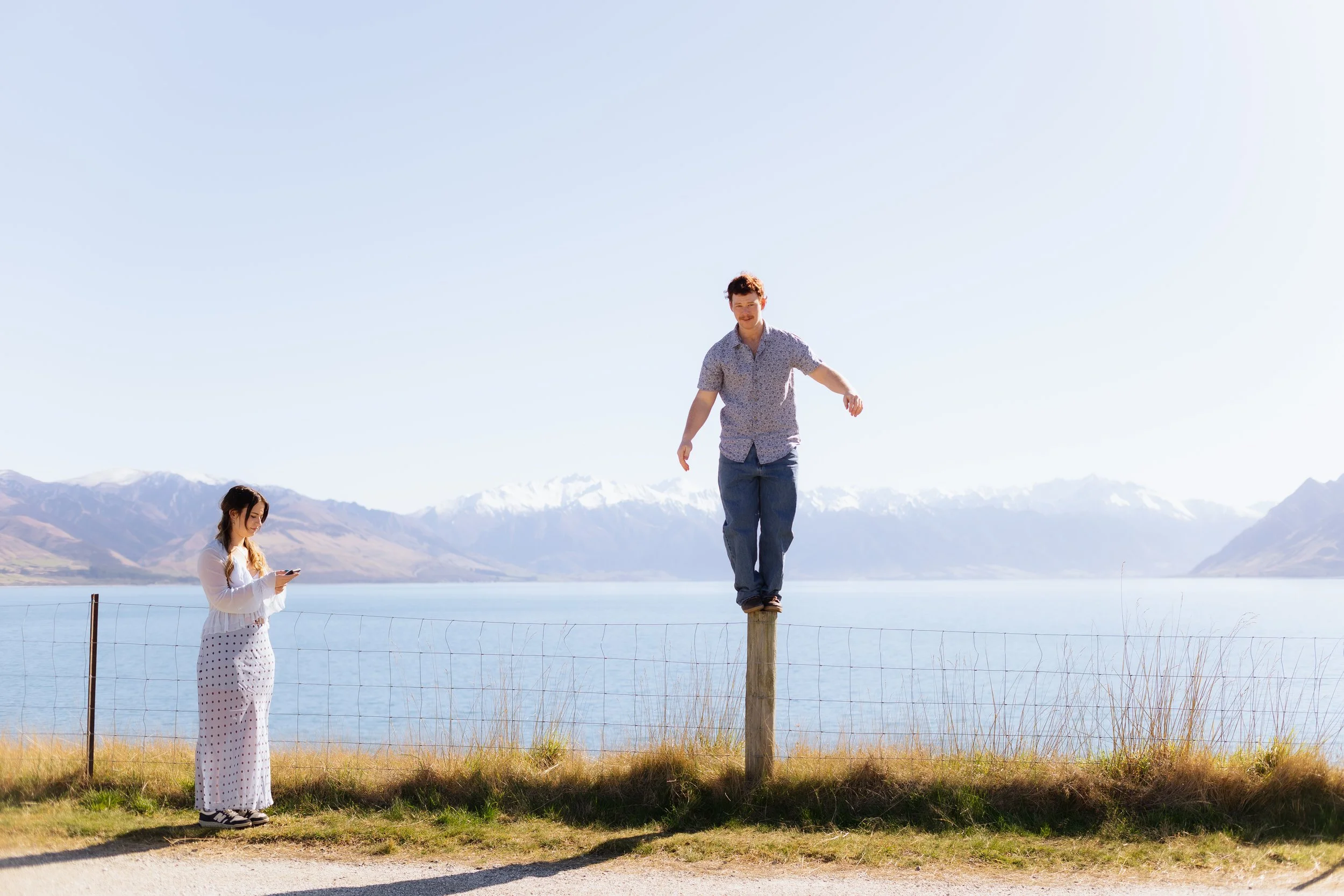 A man balancing on a wooden post while two women stand nearby outdoors near a lake with mountains in the background, under a clear blue sky.