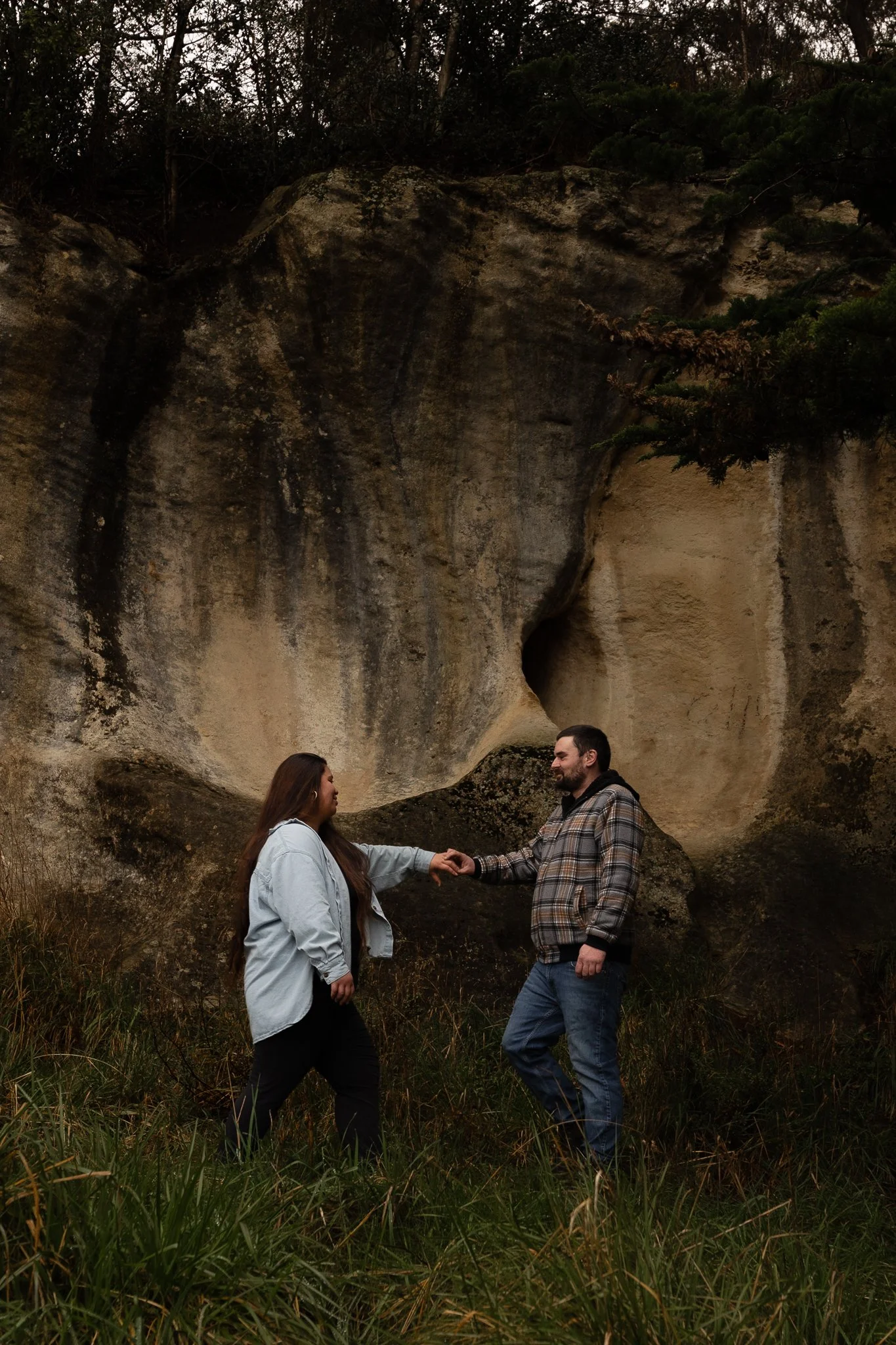 A man and a woman stand outdoors holding hands in front of a large rock formation with trees on top. The woman has long hair and wears a light-colored jacket, the man has short hair and a beard, and wears a plaid shirt. They are looking at each other