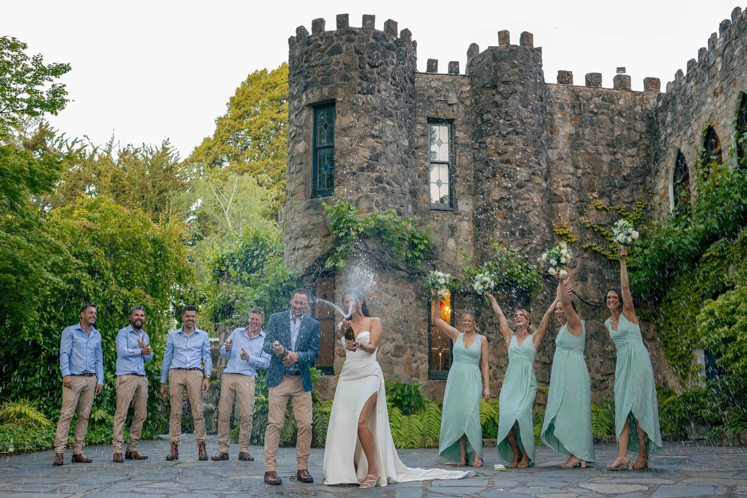 A wedding celebration outdoors with a bride and groom spraying champagne, surrounded by bridesmaids and groomsmen in front of a stone castle-like building.