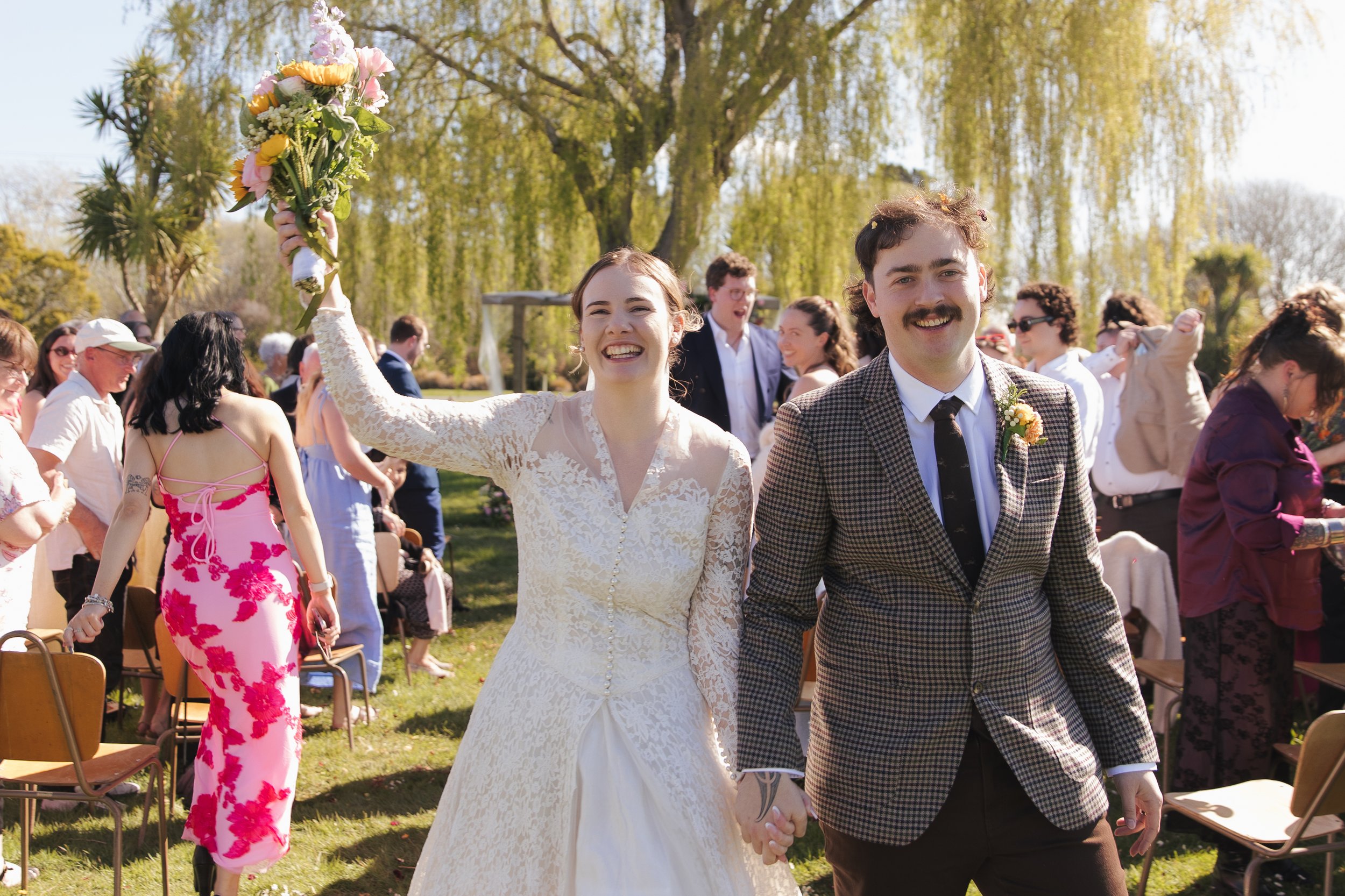 Bride wearing a vintage wedding gown is holding her bouquet in the air as she walks down the isle with her husband who is wearing a plaid suit jacket