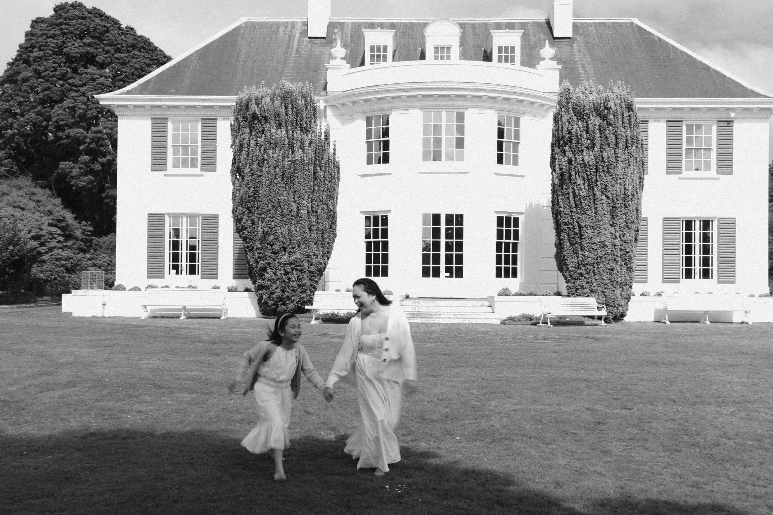 A woman and a girl holding hands and running on grass in front of a large, white, multi-story house with multiple windows, shutters, and two tall trees in the front yard, in black and white.
