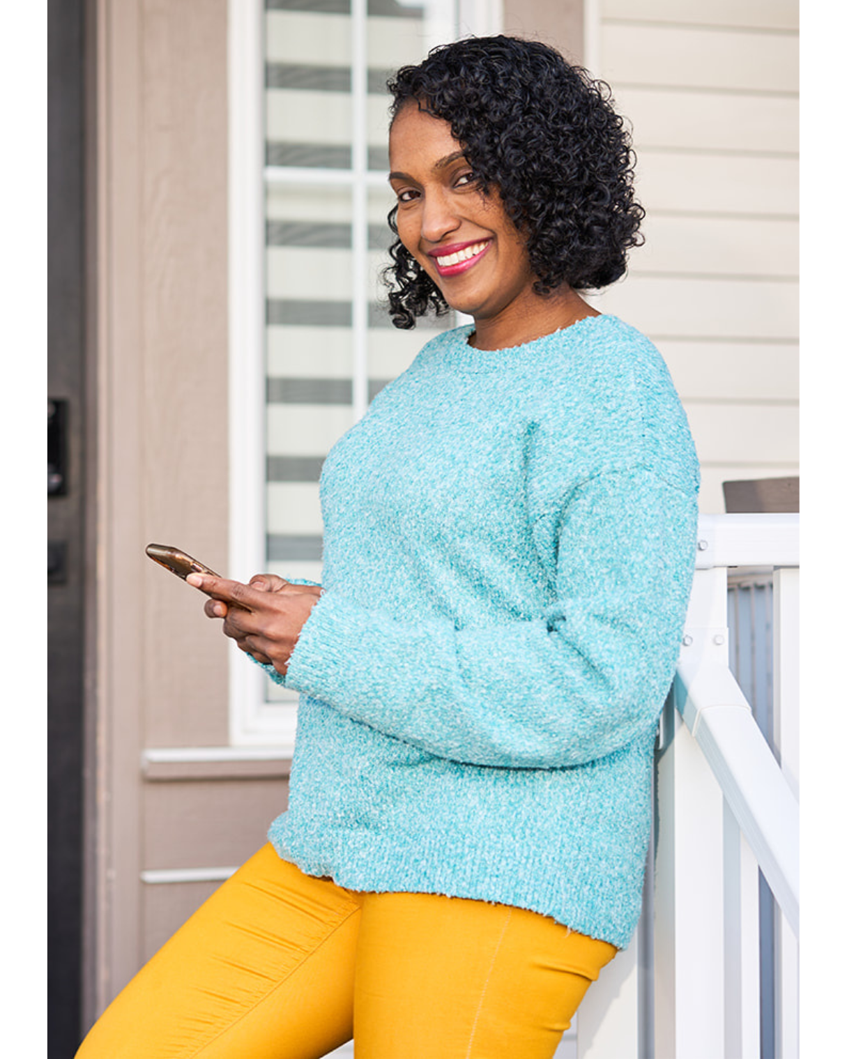 A woman with curly black hair smiling and holding a smartphone. She is standing outside on a porch, wearing a light blue sweater and yellow pants.