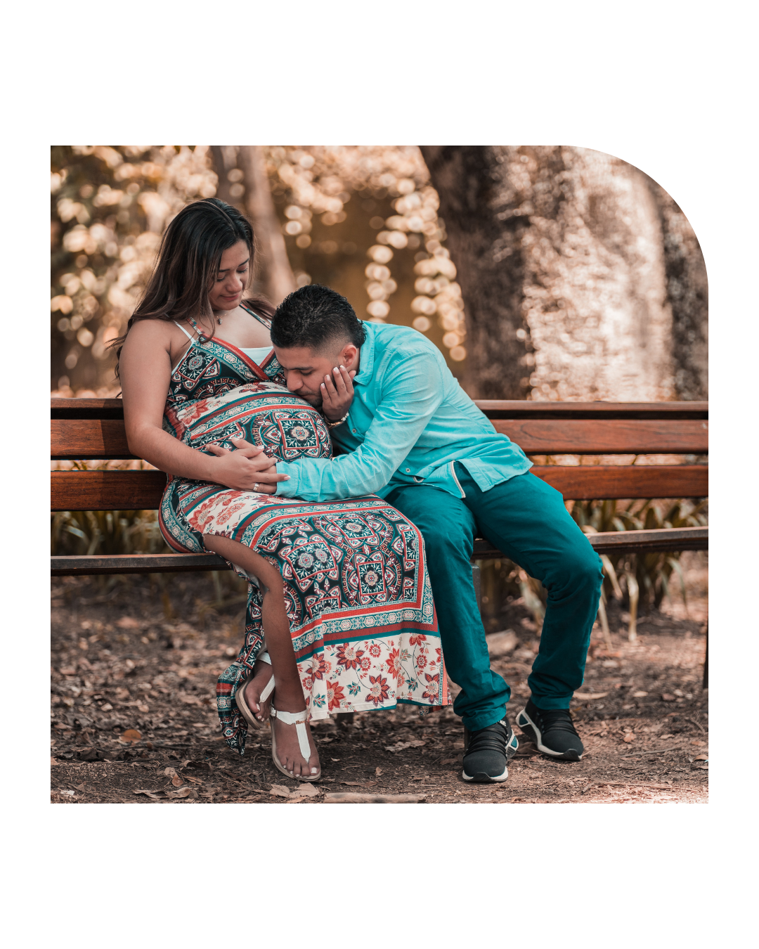 Pregnant woman sitting on a park bench with her partner, who is resting his head on her belly, in an outdoor park setting.