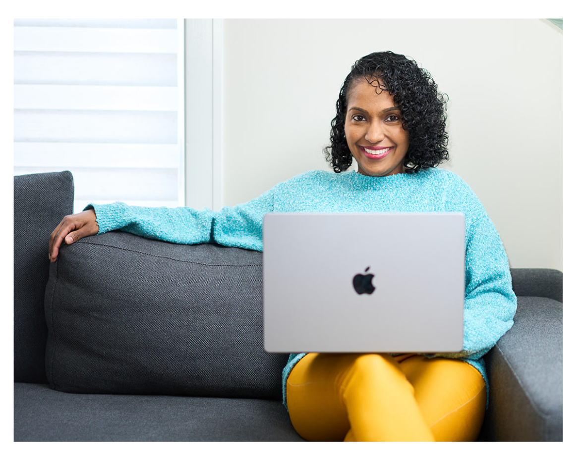 A woman sitting on a gray couch with a silver laptop on her lap, smiling.