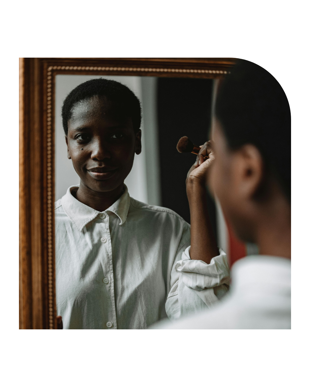 A woman applies makeup while looking in a mirror, with her reflection showing her face and the makeup brush in her hand.