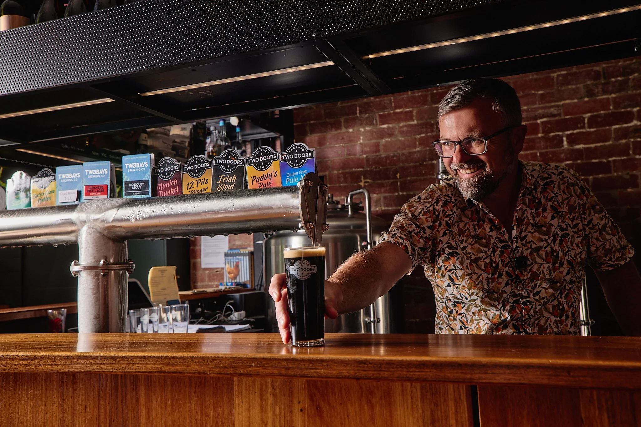 A man serving a dark beer in a pint glass at a bar with a brick wall background and beer signs.